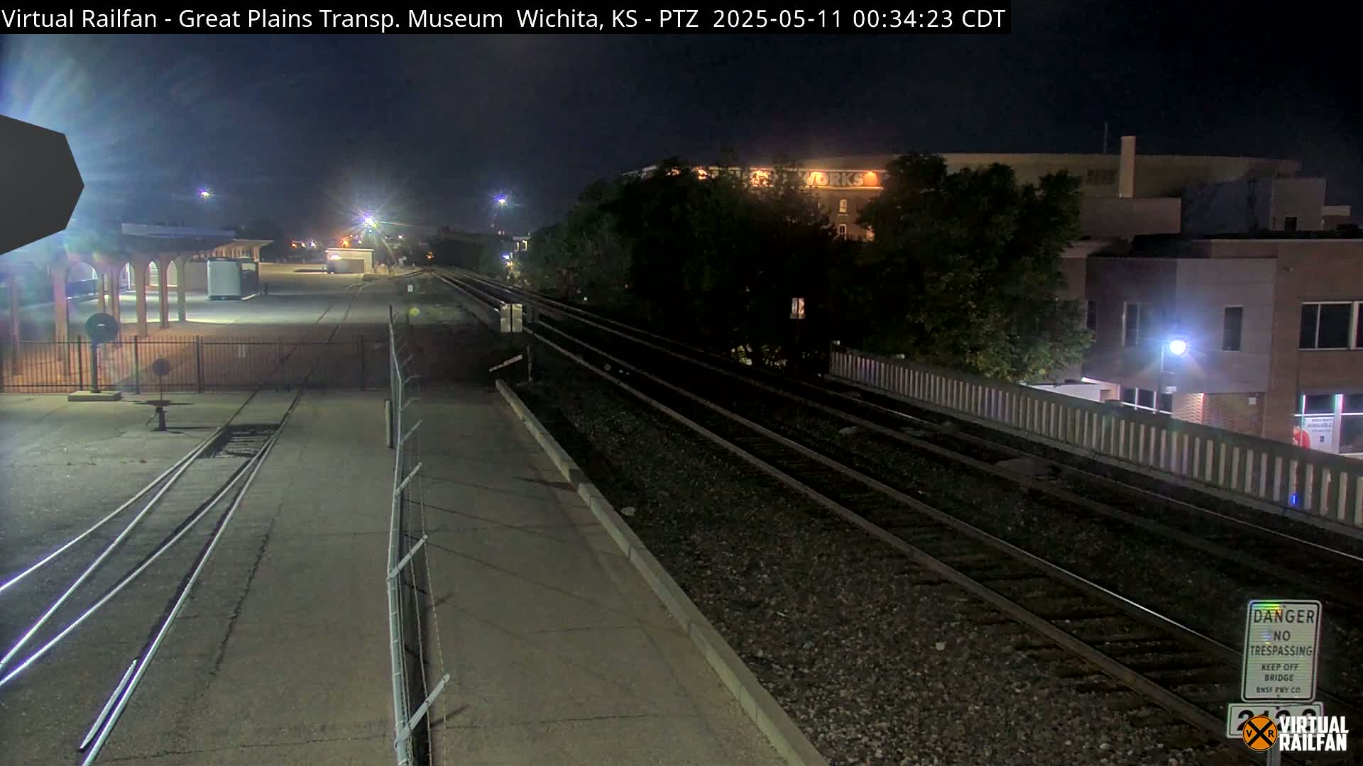 At night, a train track stretches into the distance, passing by a fenced-in area and a building.