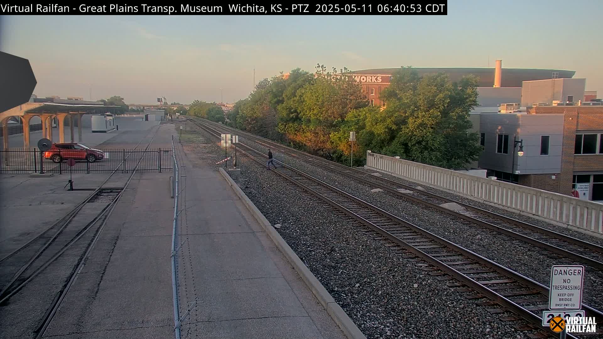 A person walks along railroad tracks under a mostly clear, sunrise sky, with a car parked nearby and buildings visible in the distance.
