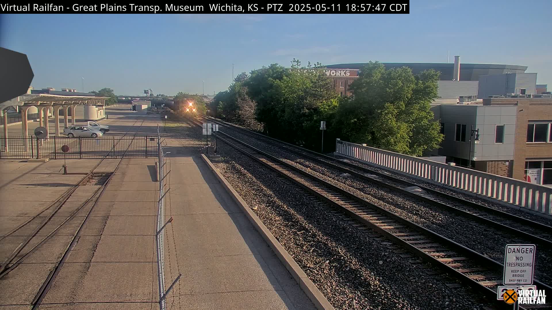 A train approaches a railway crossing under a clear, sunny sky, passing by a station and various buildings.