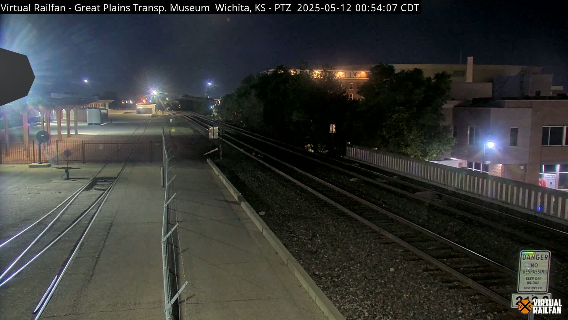 Nighttime view of train tracks at a station, with a building and trees in the background.