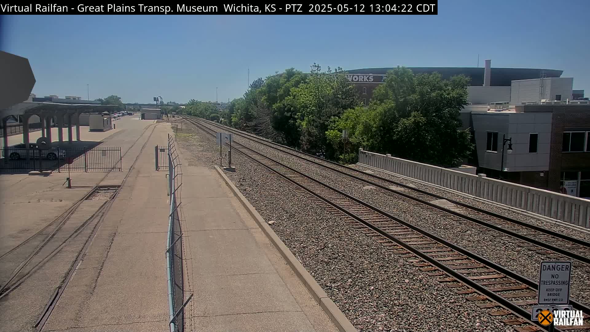 A sunny day reveals train tracks extending into the distance, passing a paved walkway and a covered platform before reaching a low wall and buildings beyond.