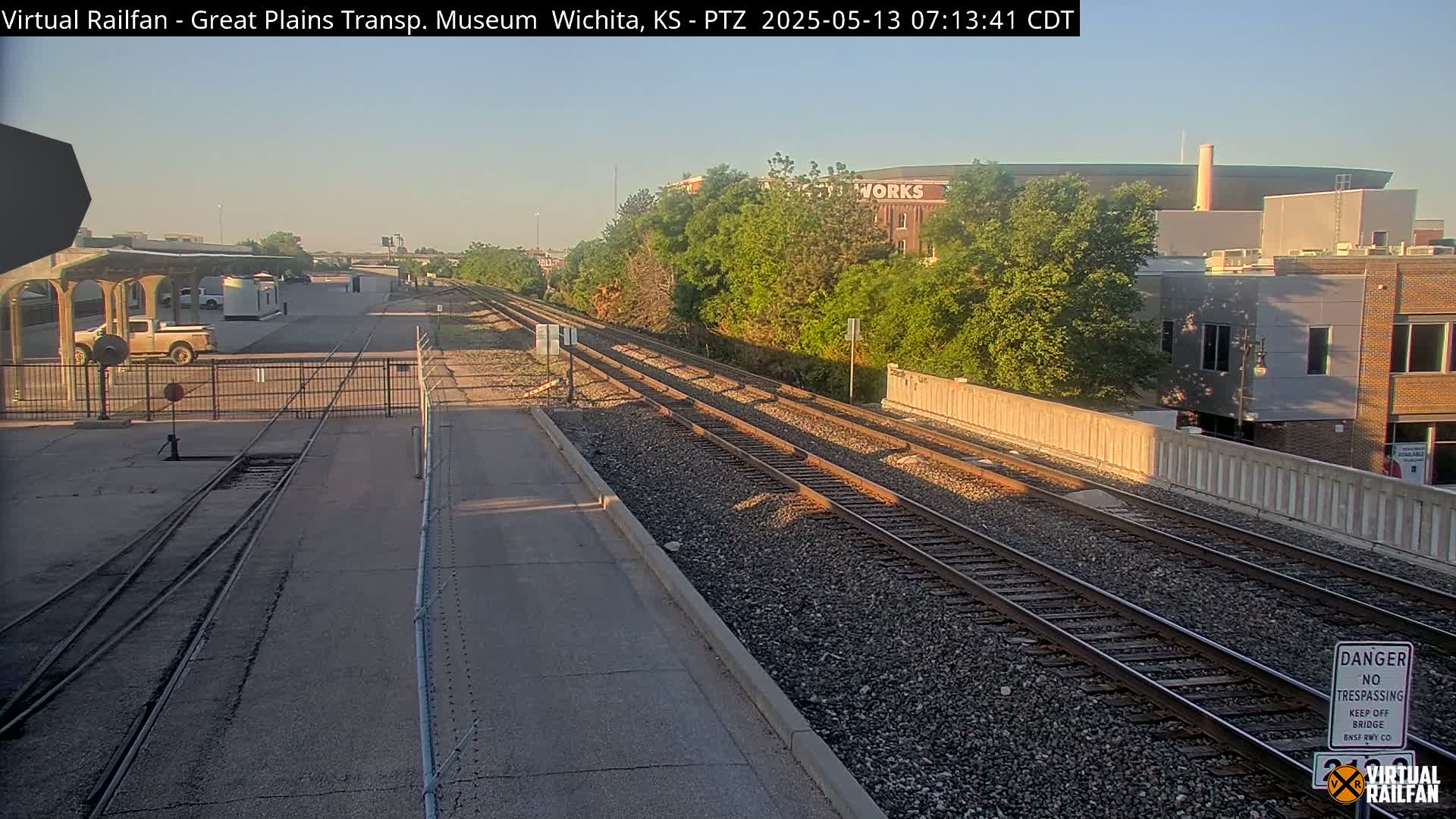 A sunny view of a train station with multiple tracks, a concrete platform, and a low fence, surrounded by greenery and buildings in the background.