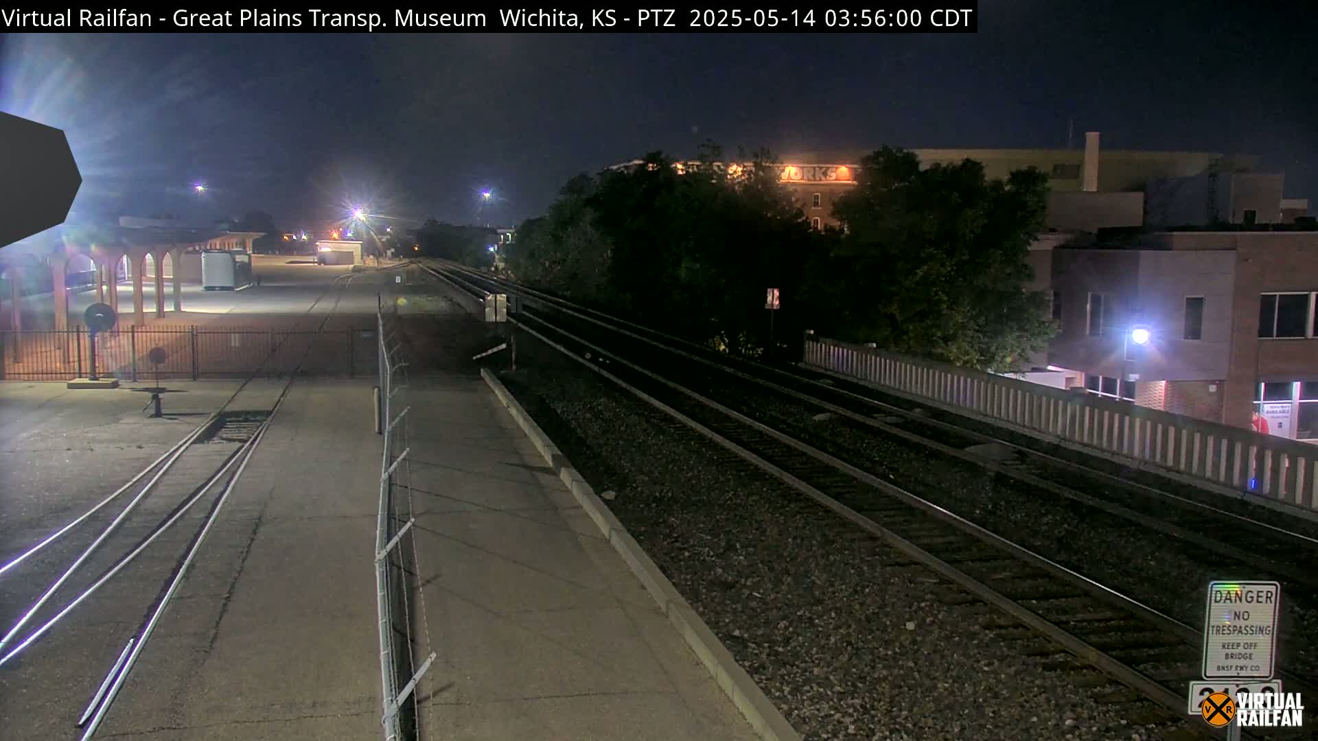 A nighttime view of train tracks beside a building and a fenced-off area, under a dark sky.