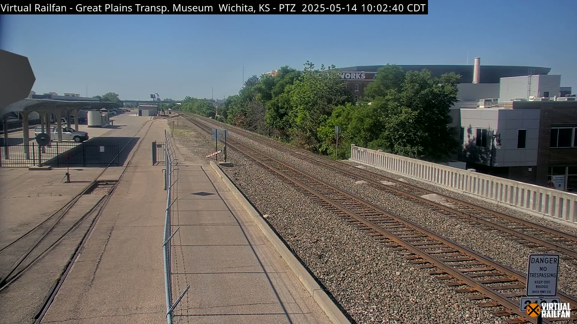 A sunny day reveals a railway track flanked by a paved road and a low concrete wall, leading toward buildings and lush greenery.