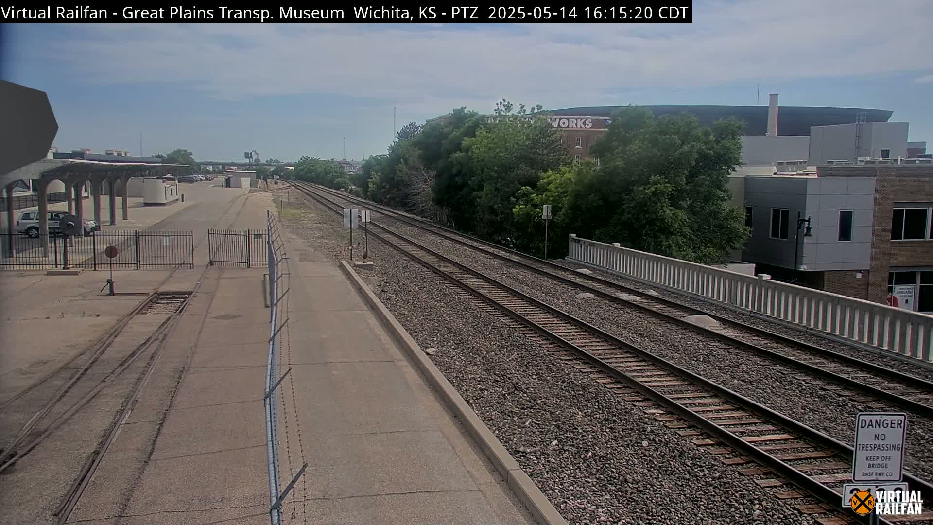 A mostly sunny view of train tracks extending into the distance, passing by a fenced-in area and several buildings, under a mostly clear sky.