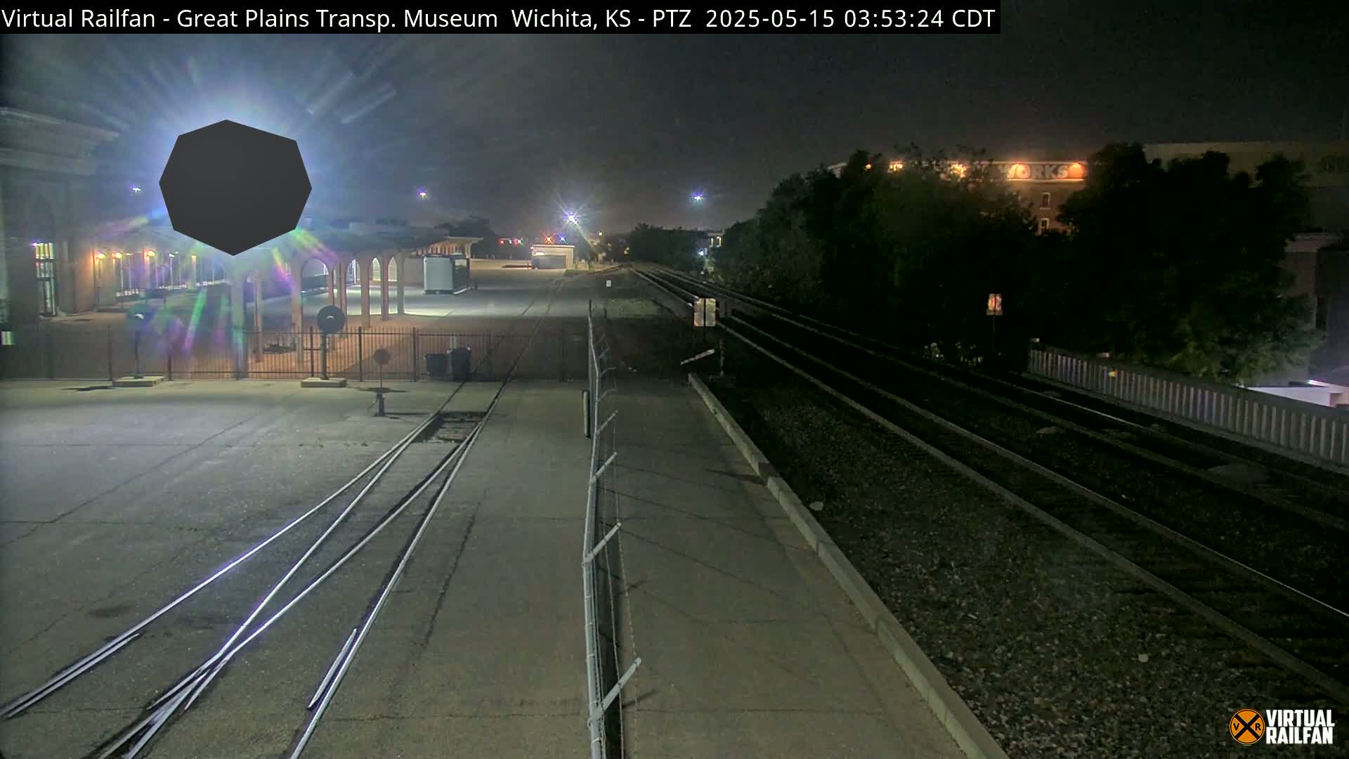 A nighttime view of a railway yard with converging train tracks, a building, and a large, dark, polygon-shaped object obscuring a section of the scene.