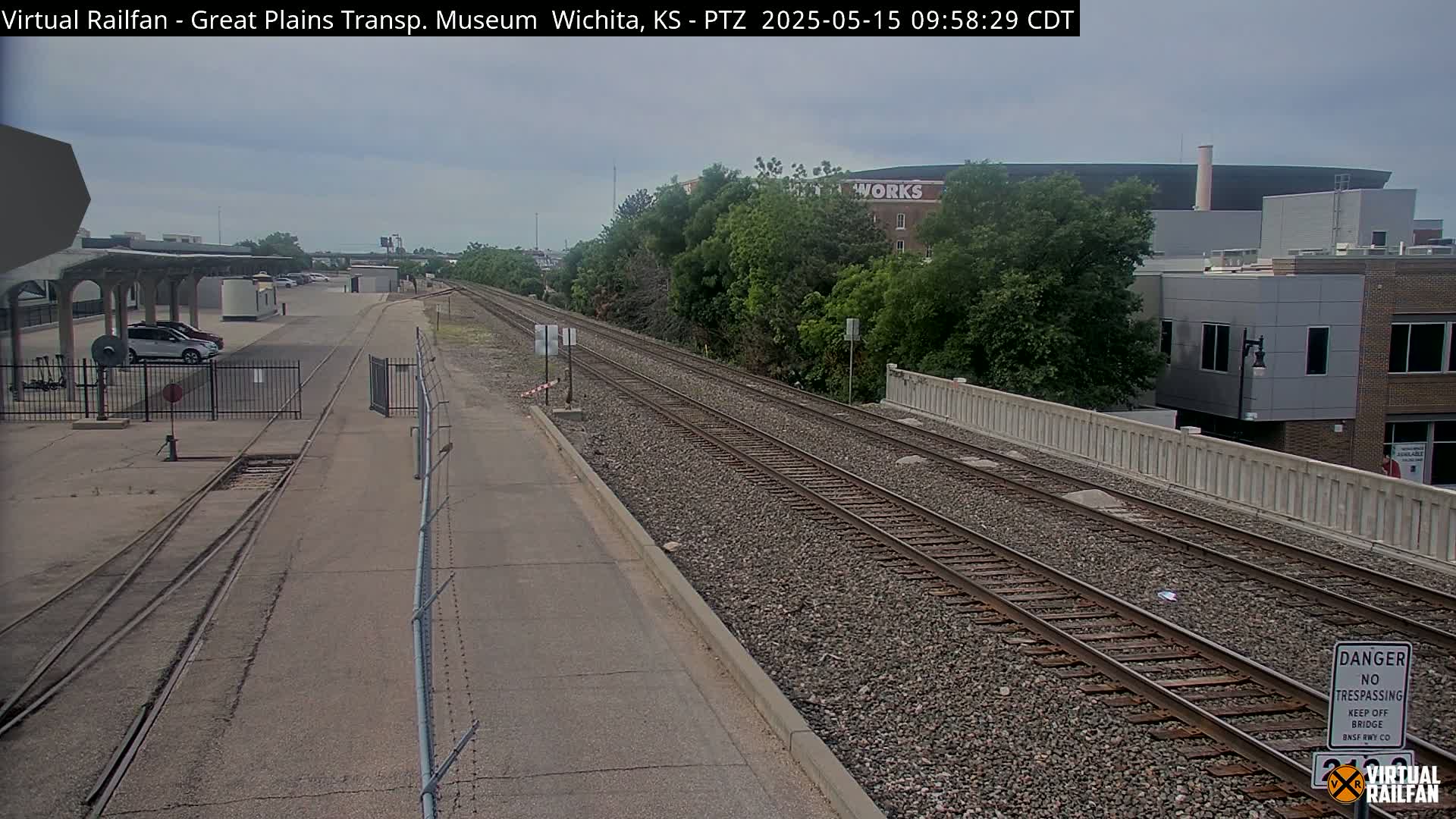 A mostly cloudy day shows two sets of train tracks alongside a paved road, with a parking lot and buildings visible in the background.