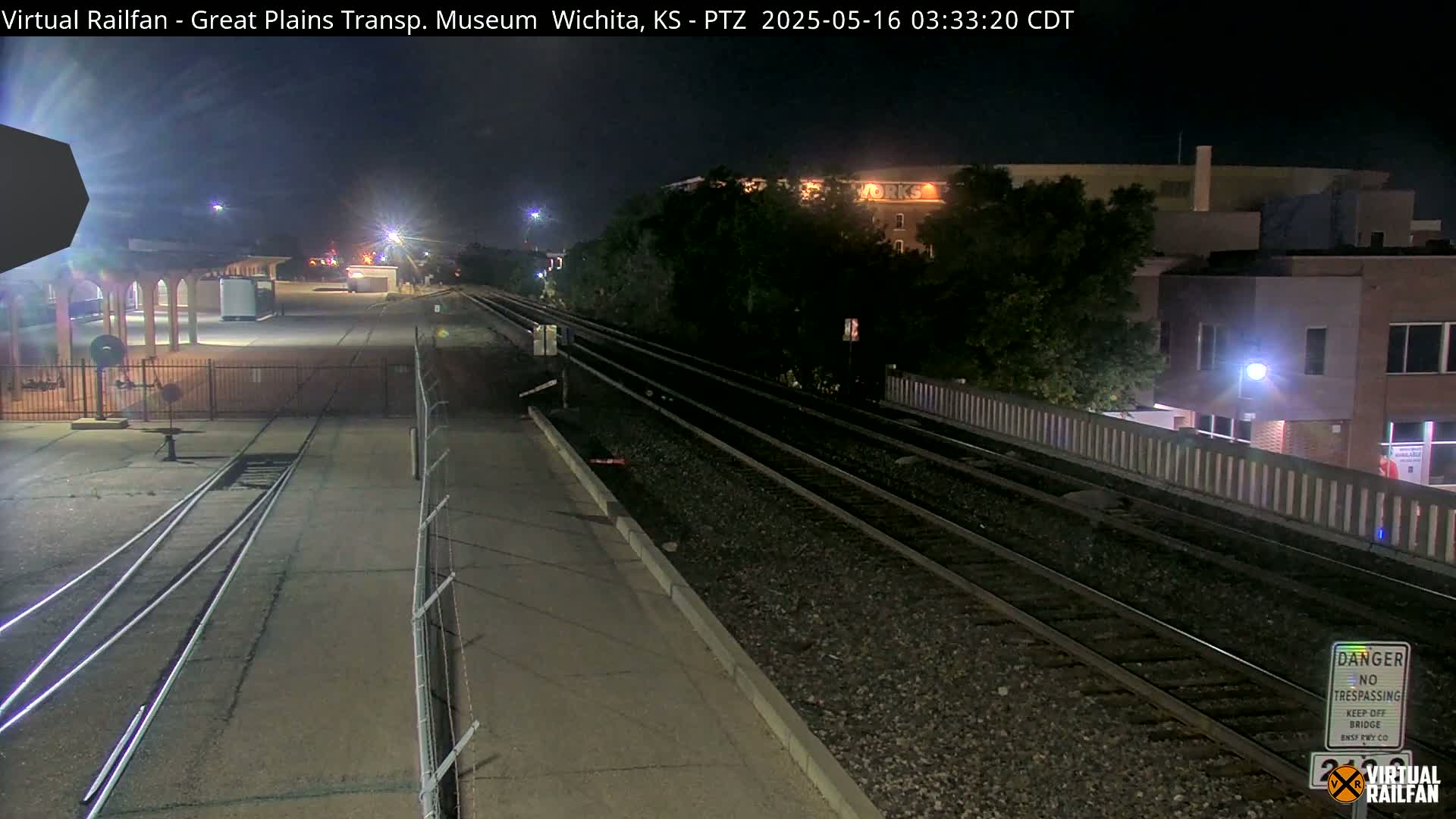 At night, a train track runs through a mostly empty station area near some buildings, under partly cloudy skies.