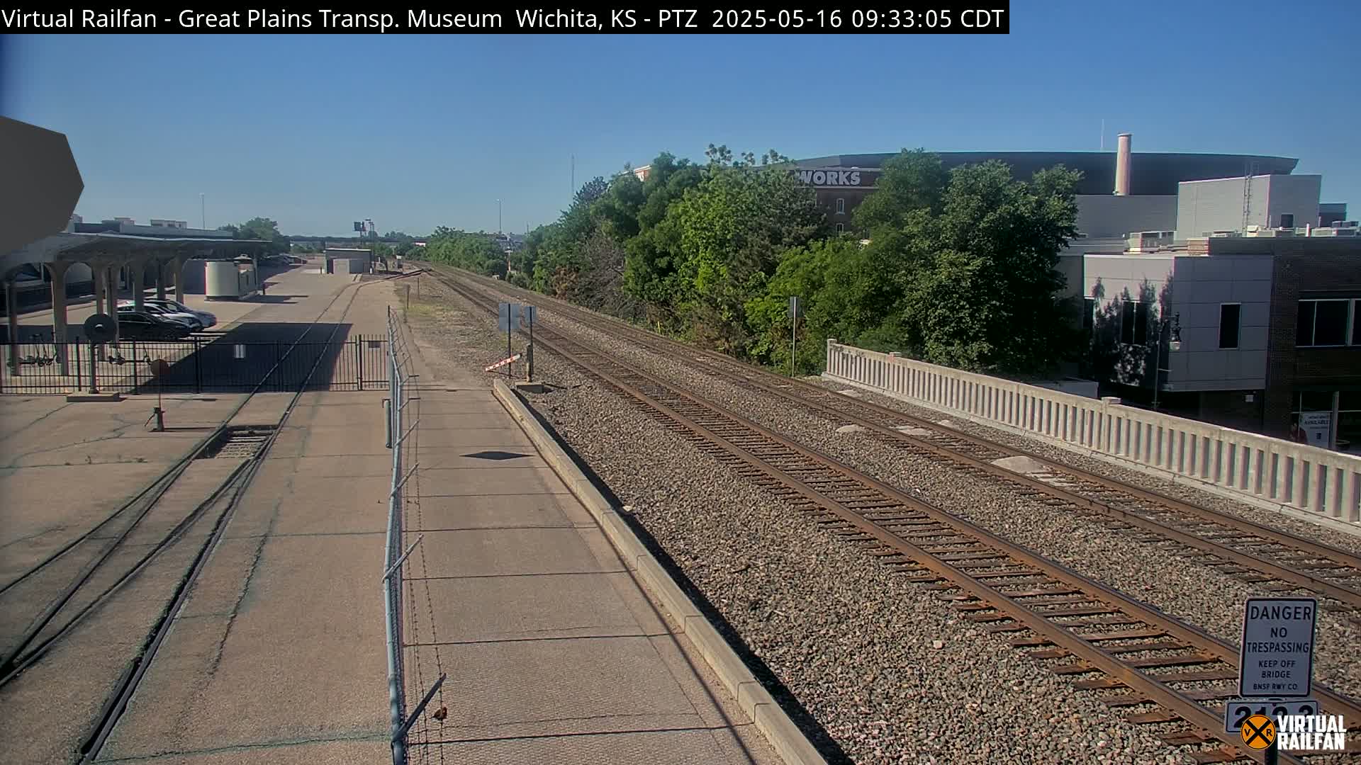 A sunny view of railroad tracks extending into the distance, passing alongside a low wall and a building, with several parked cars visible in a nearby lot.