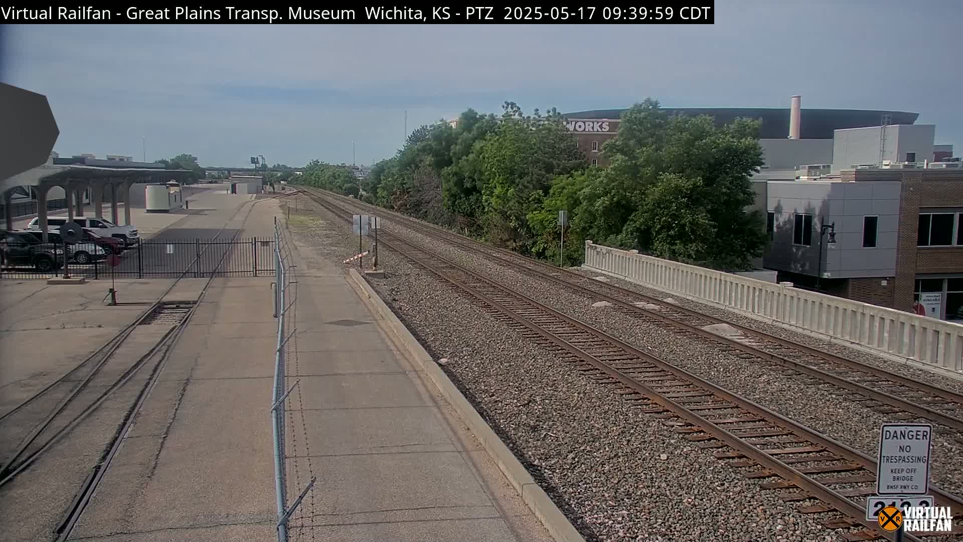 A view of railroad tracks extending into the distance under a partly sunny sky, with cars parked in a lot to the left and buildings visible in the background.