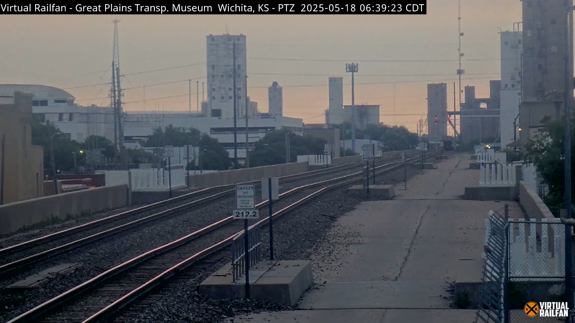 A view of railroad tracks extending into the distance under a hazy, sunrise sky, with industrial buildings and structures visible in the background.
