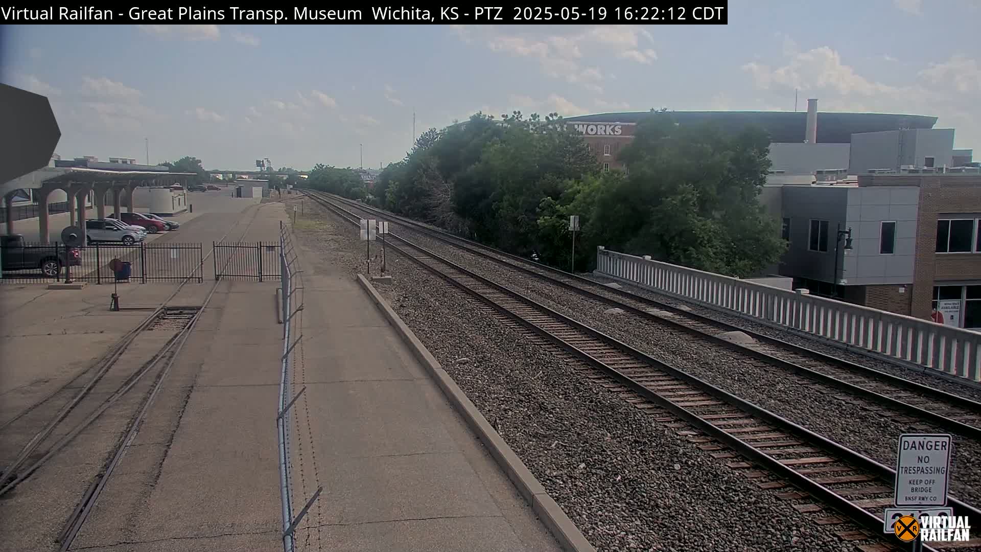 A view of railroad tracks stretching towards a bridge and buildings under a partly cloudy sky.