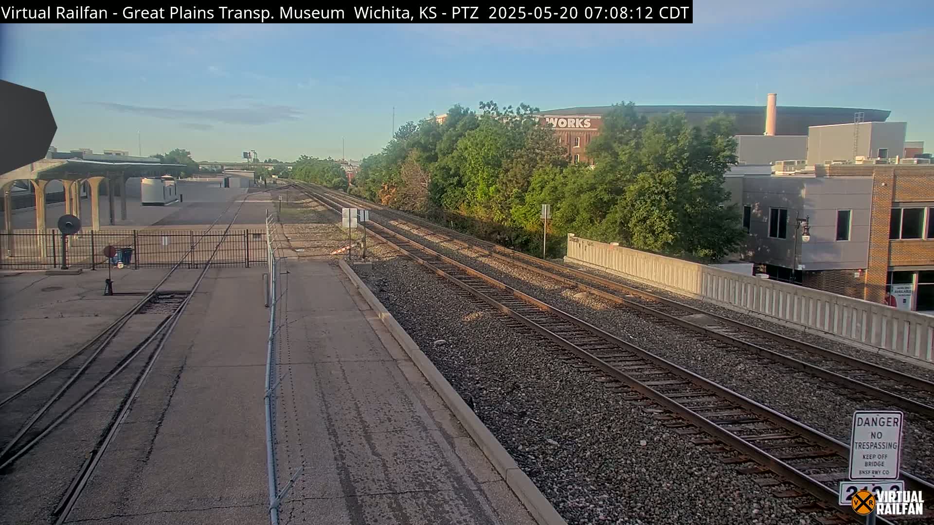 A sunny morning view of railroad tracks extending into the distance, alongside a paved road and bordered by buildings and lush green trees.