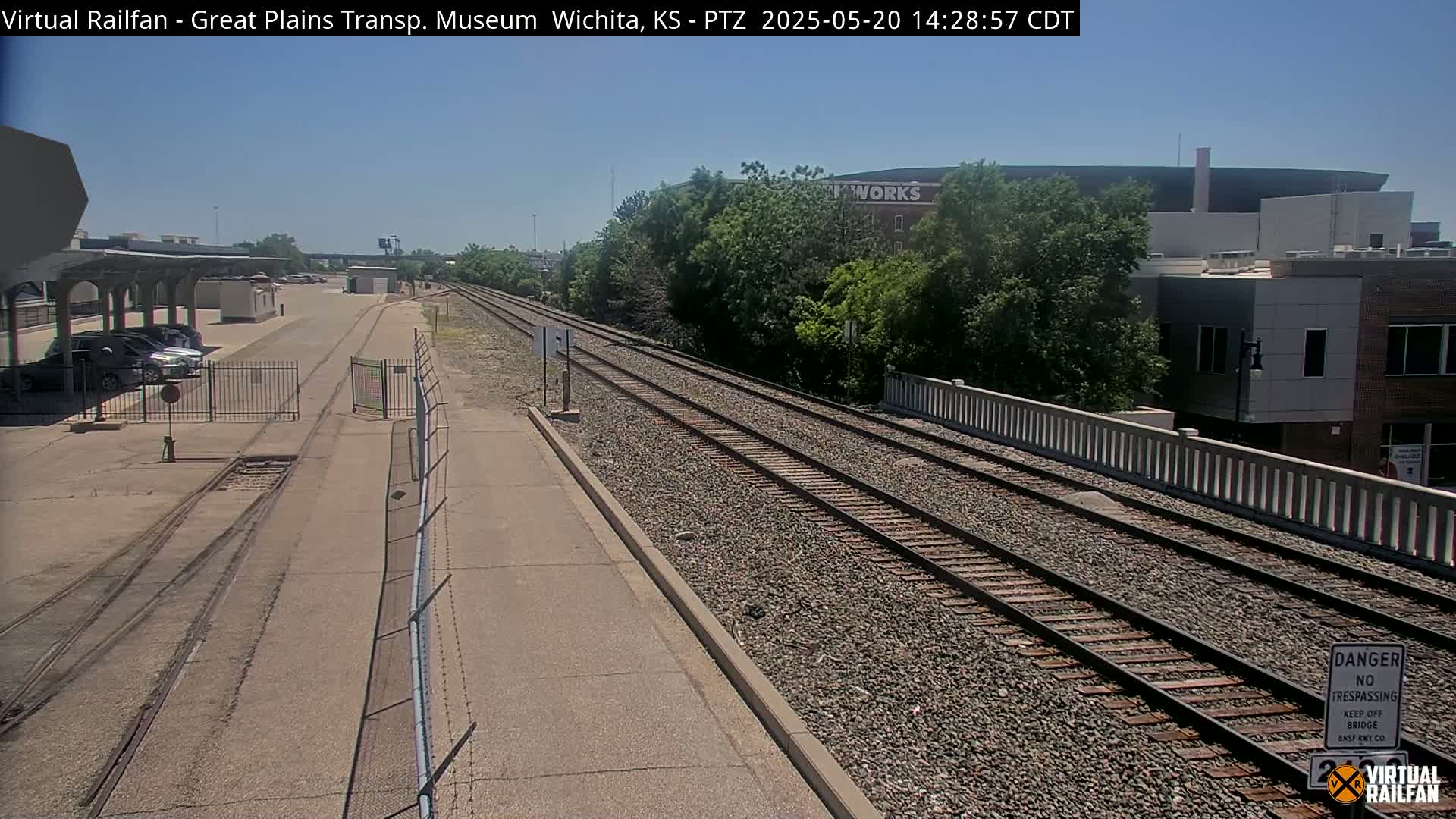 A paved area next to railroad tracks on a sunny day, with parked cars visible in the distance and buildings beyond the tracks.