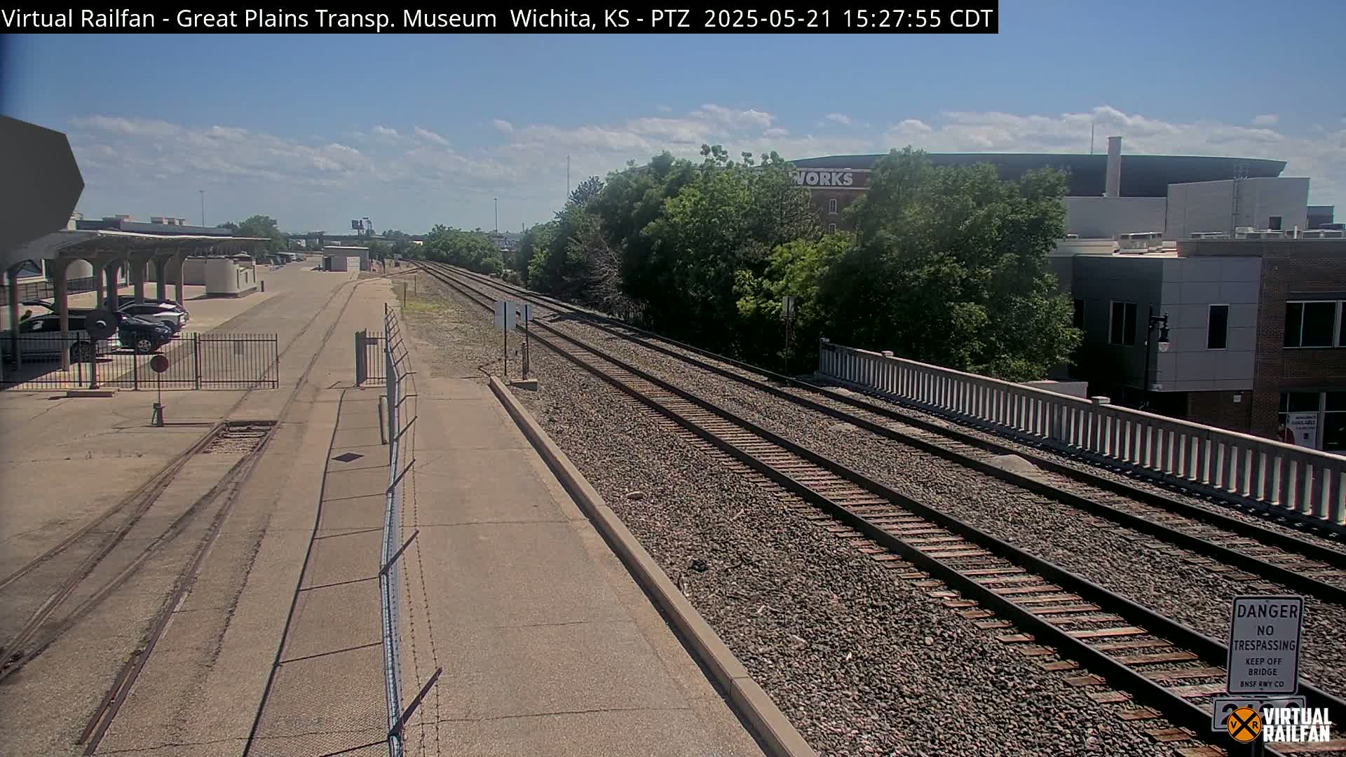 A sunny day reveals a train track extending from a parking lot with several cars under a covered area, passing through a fenced-off area, and toward buildings beyond a bridge.