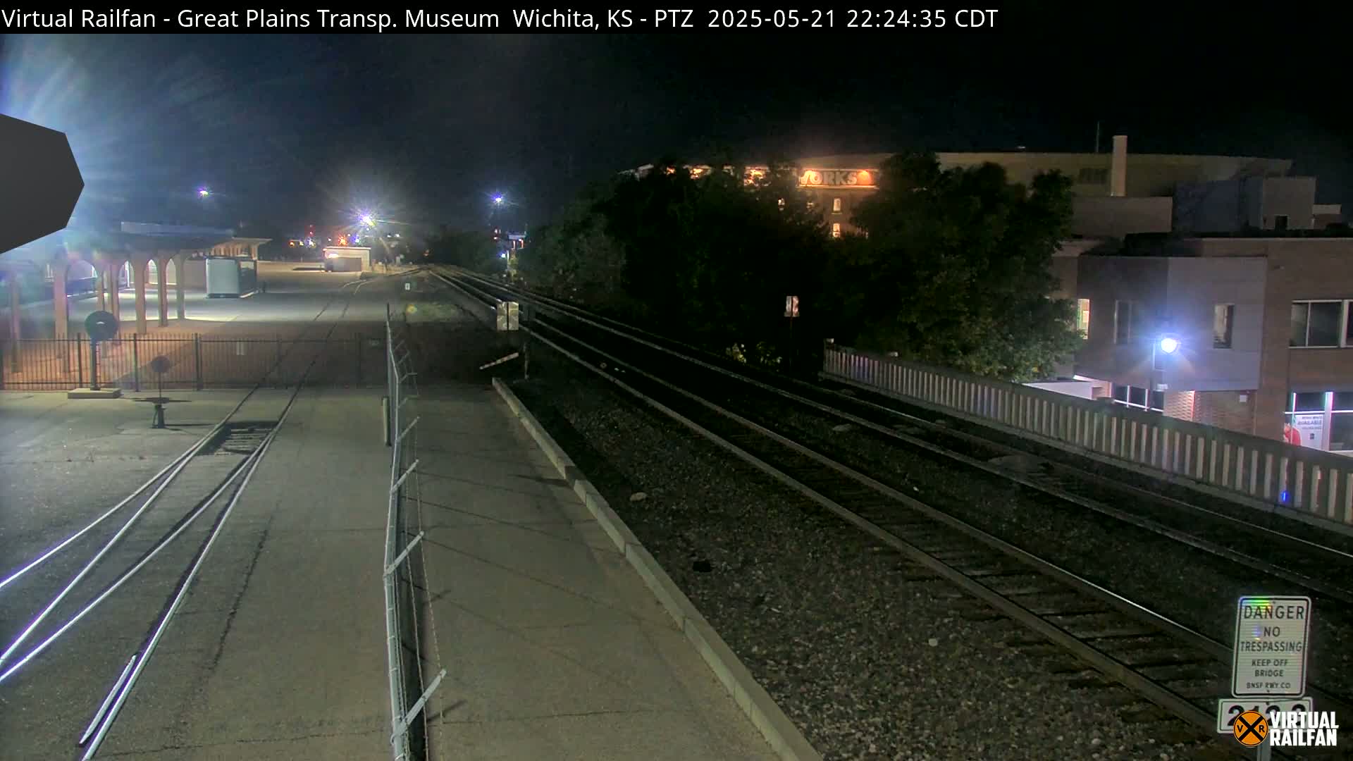 At night, railroad tracks extend into the distance, passing by a low brick building and a fenced-in area with a covered structure.