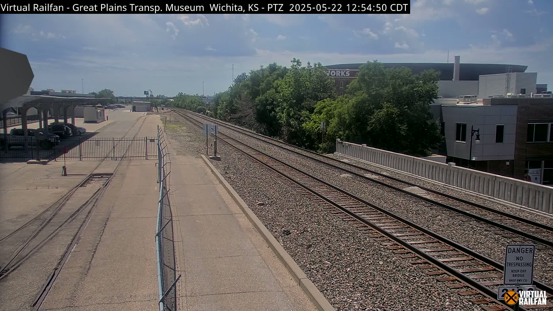 A sunny day view of railroad tracks extending from a fenced-in area with parked cars, past a low wall, towards buildings and trees in the distance.