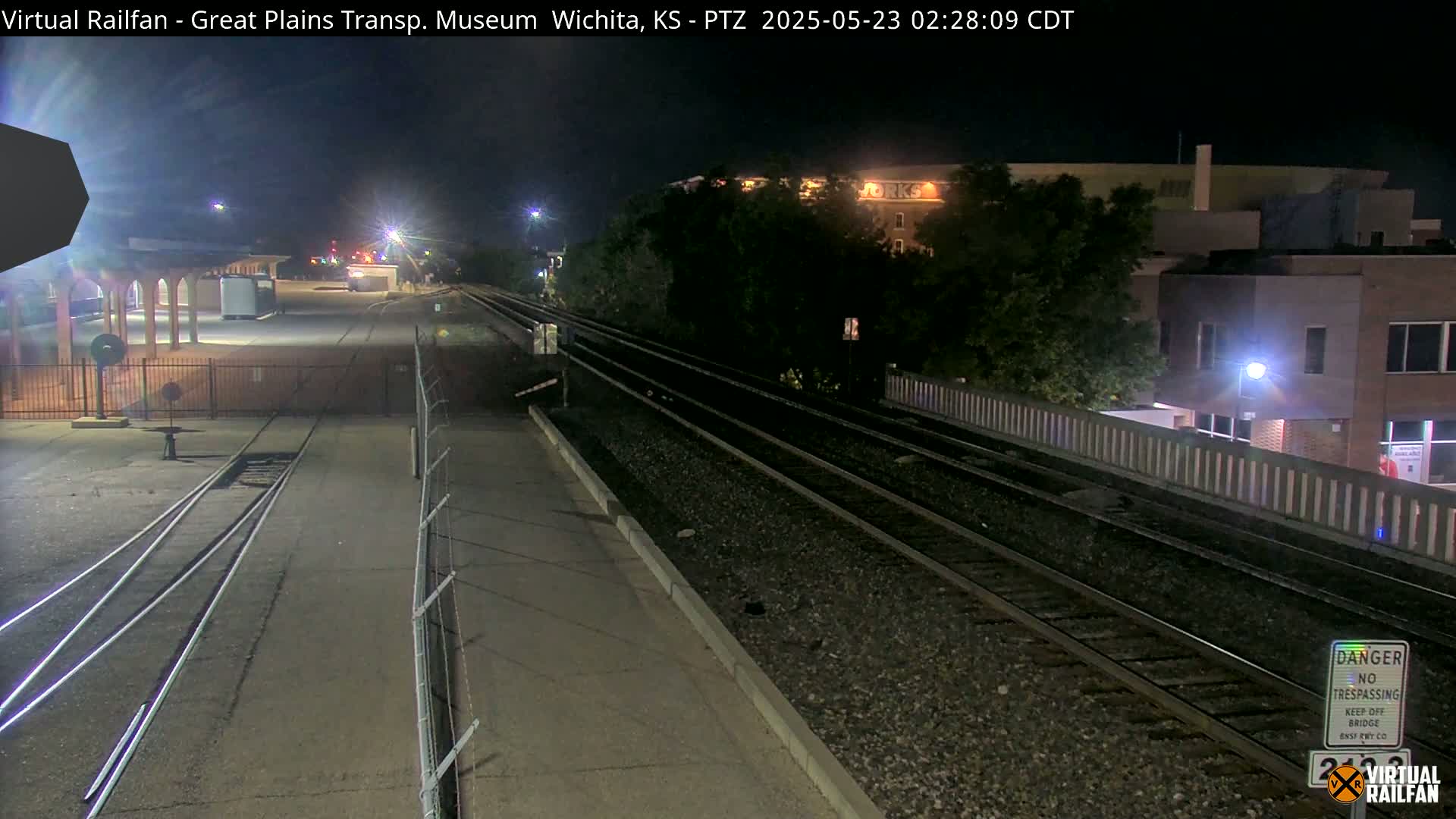 A nighttime view of train tracks and a railway station platform, with buildings and trees in the background under dark skies.
