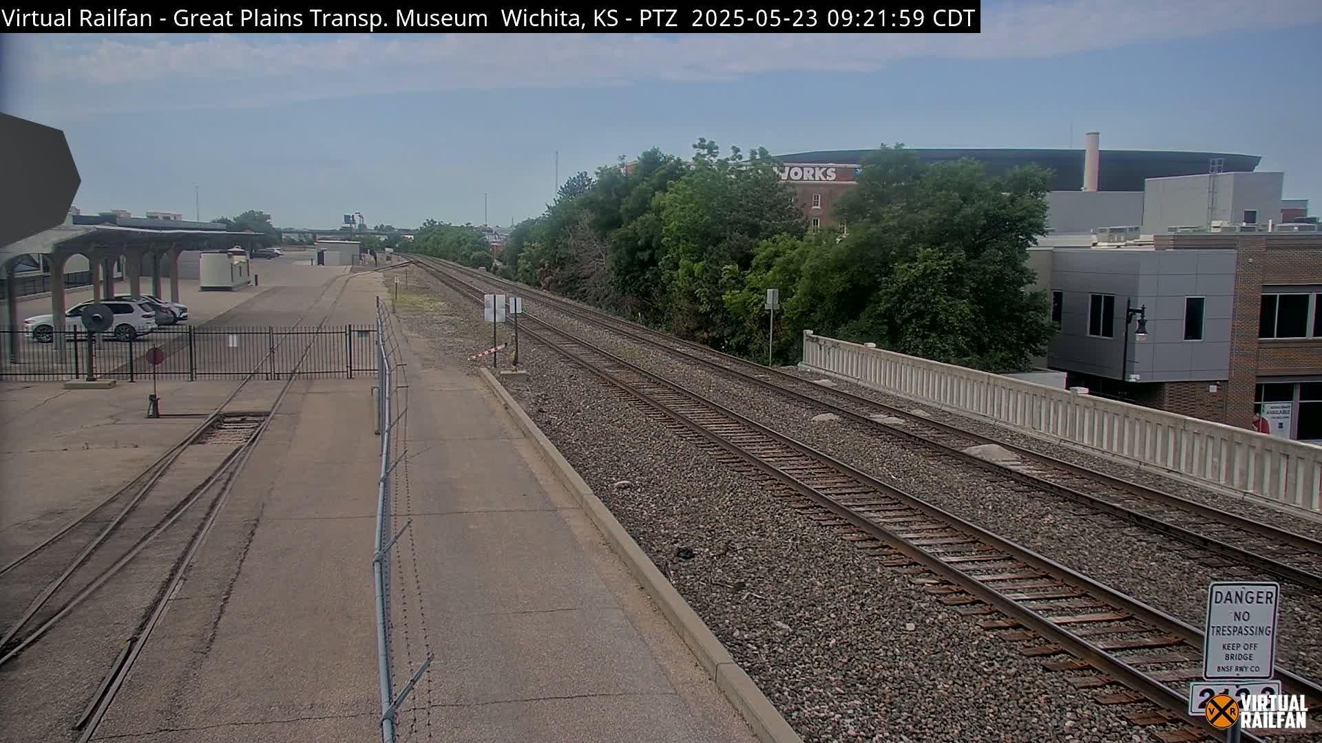 A view of railroad tracks on a partly sunny day, with a parking lot, some buildings, and lush green trees in the background.