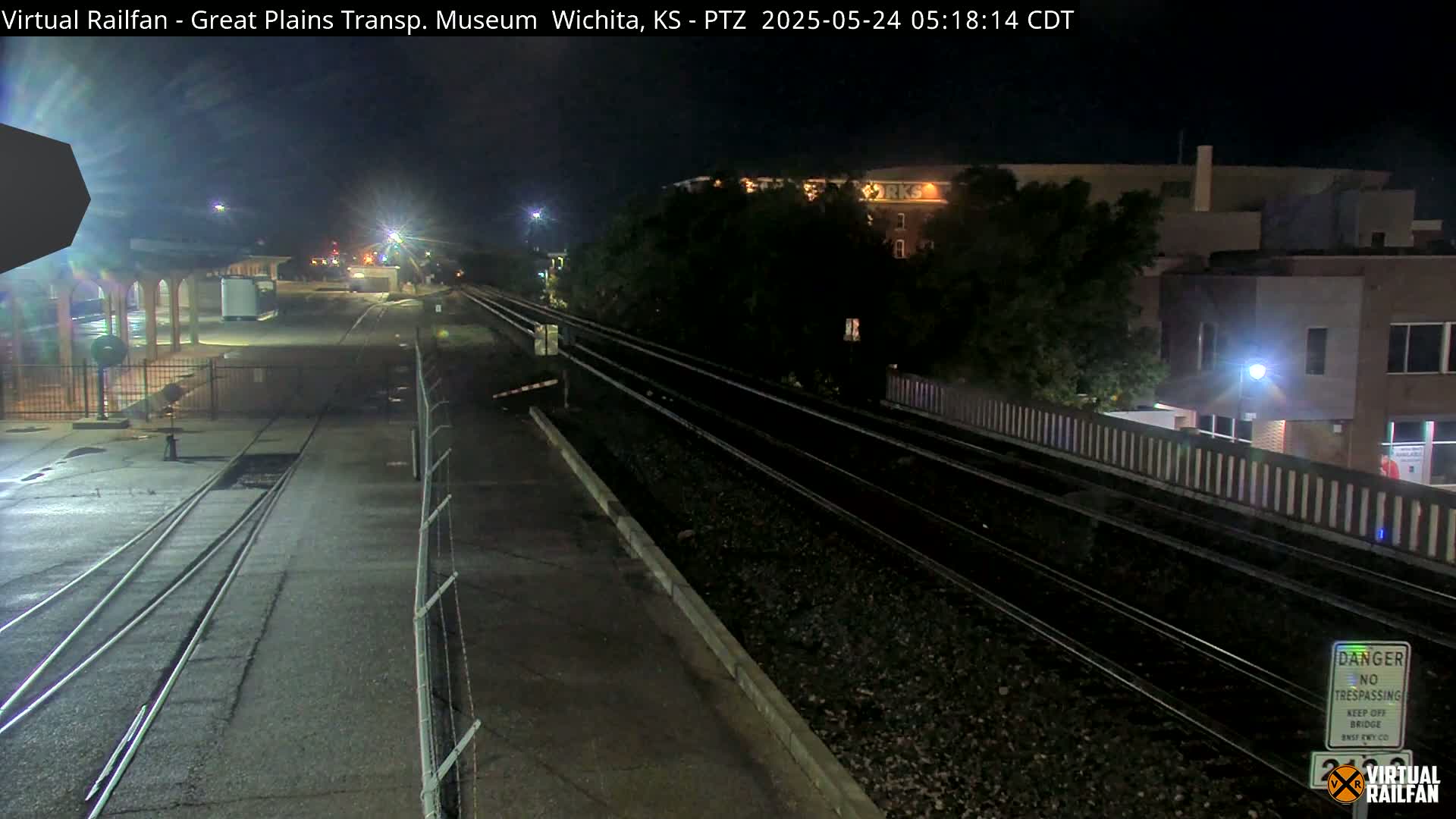At night, train tracks extend from a paved area toward a building partially obscured by trees under a clear, dark sky.