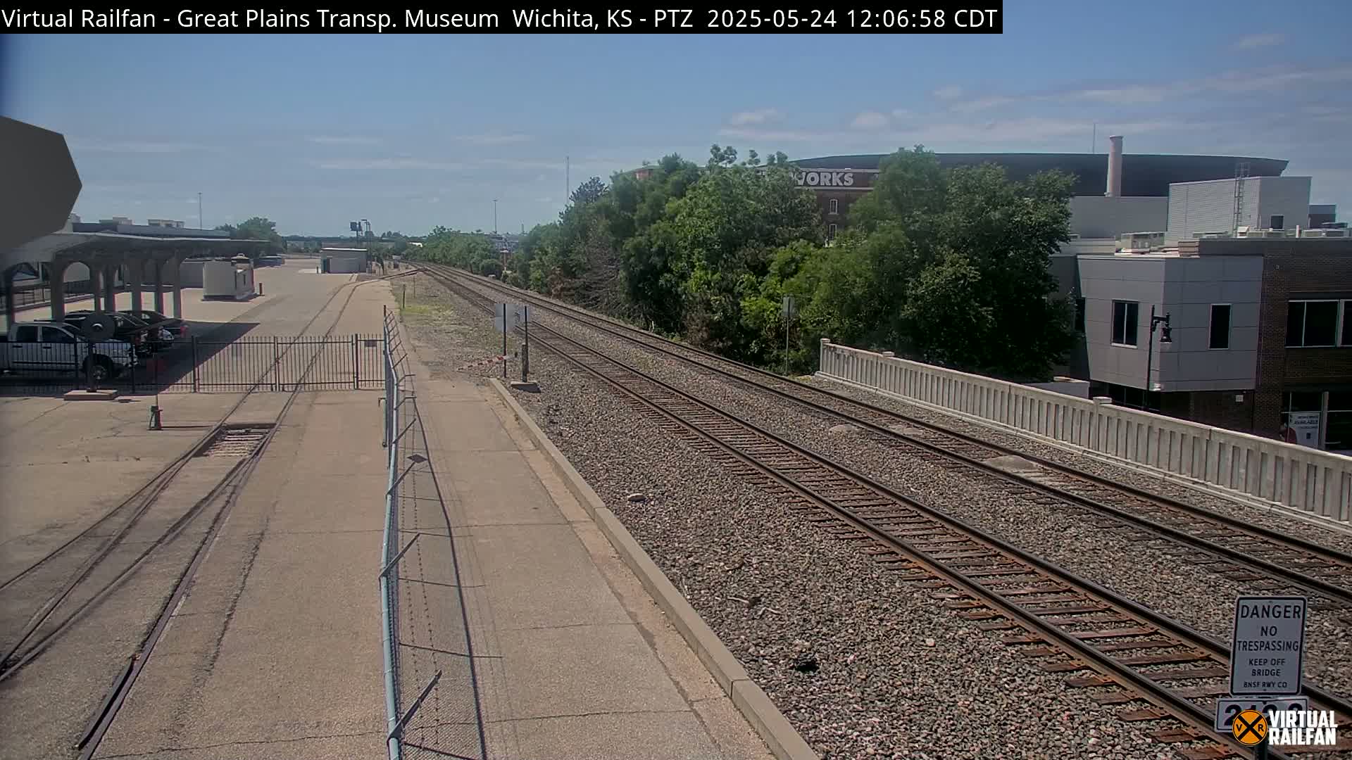 A sunny outdoor view of railroad tracks extending into the distance, passing alongside a paved area, a chain-link fence, and buildings.