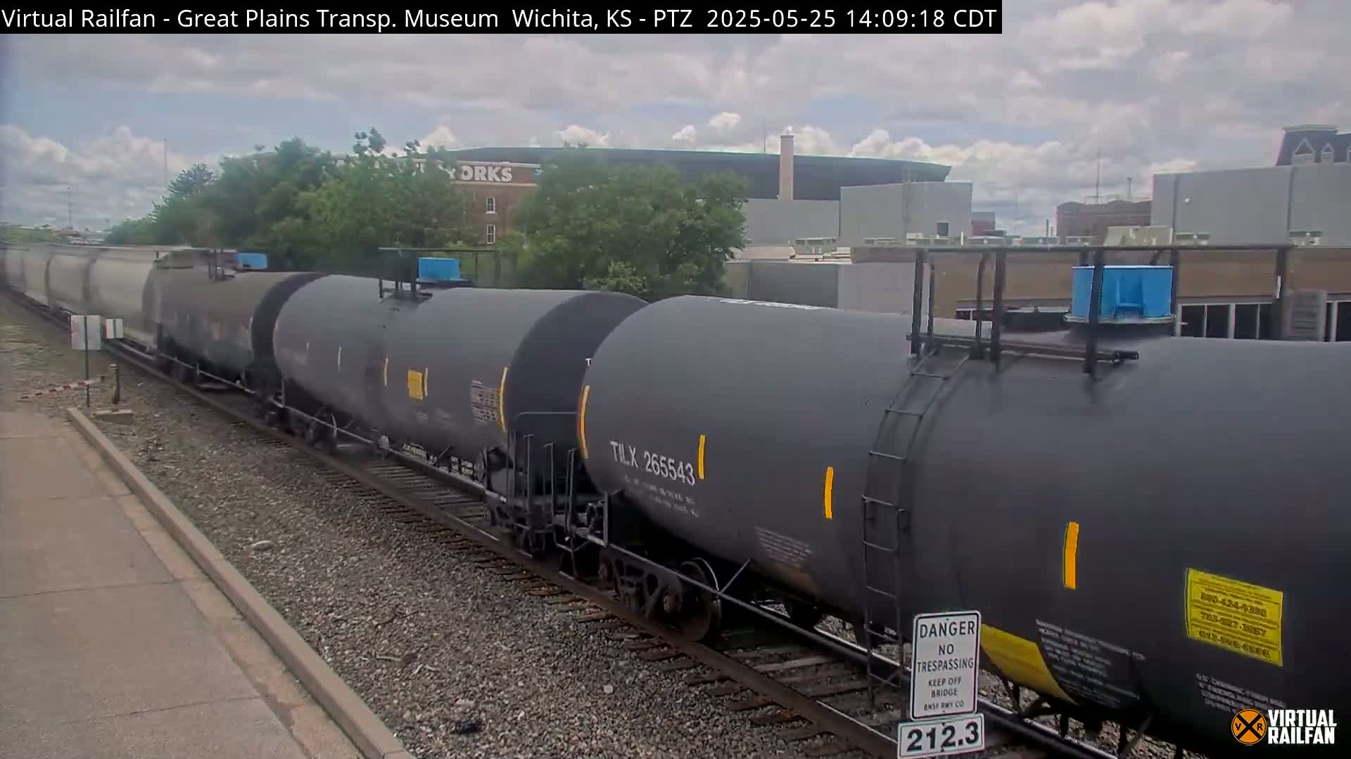 A long train of dark gray tanker cars moves along railroad tracks under a partly cloudy sky.