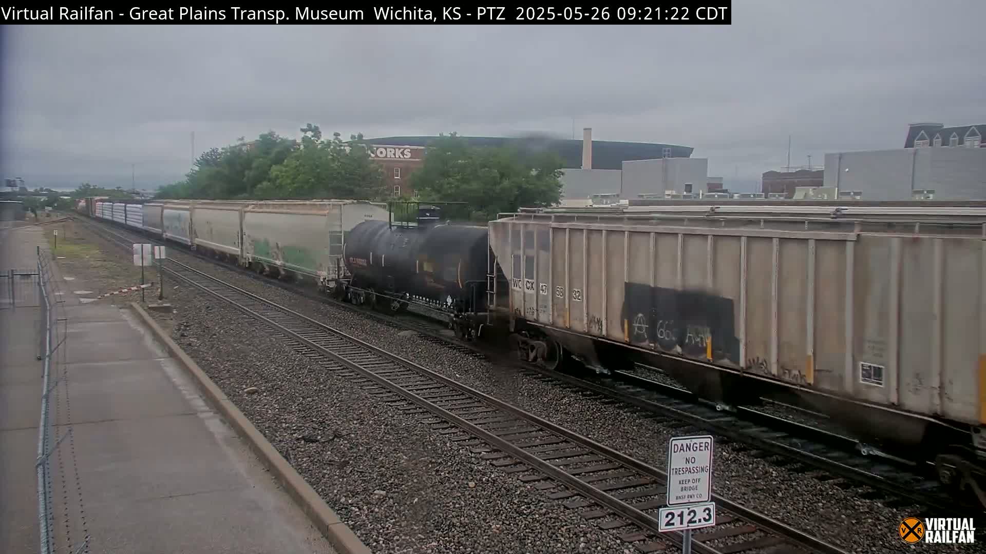 A long freight train, consisting of various cargo cars including a tanker, moves along railroad tracks under an overcast sky.