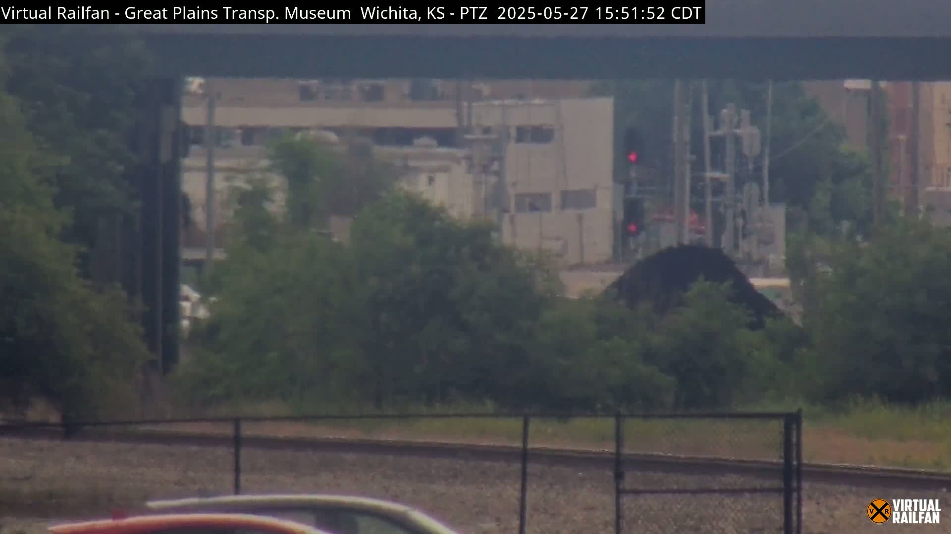 A hazy view of a train track with a dark pile of material partially obscured by greenery, in front of buildings and a red traffic signal.