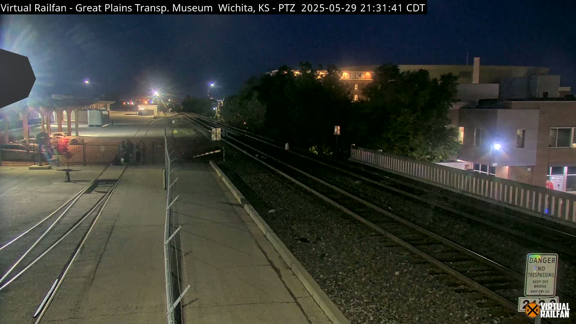 At night, train tracks extend from a paved area near a building and a chain-link fence, under a dark sky.
