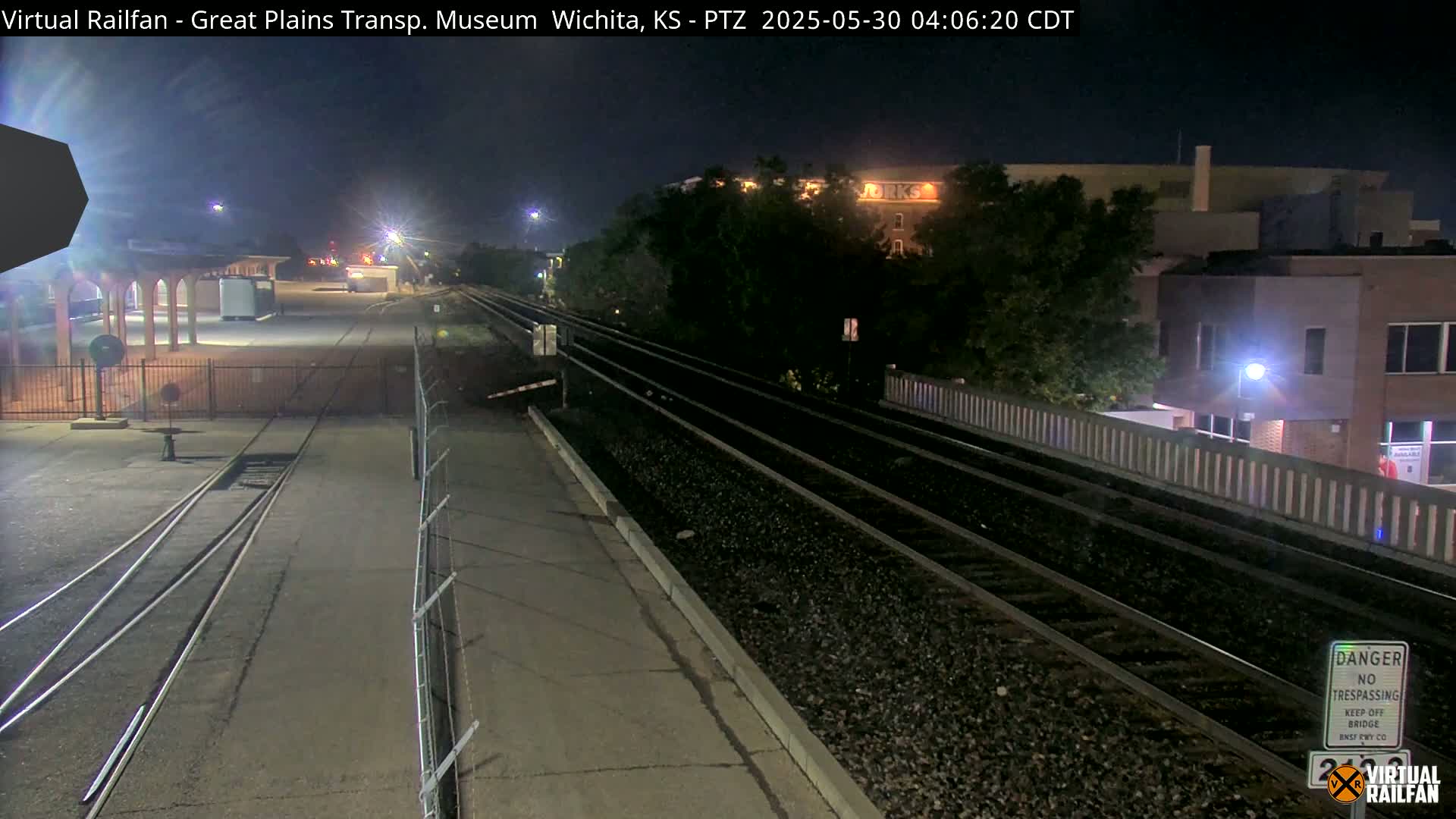 Nighttime view of railroad tracks extending into the distance, passing by a building and a fenced-in area under lightly lit conditions.