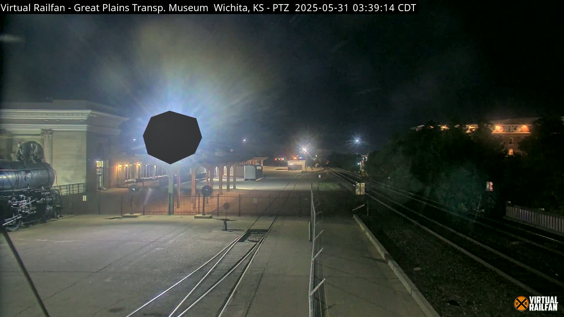 A nighttime view of a train station with a steam locomotive, railroad tracks, and a large, dark, octagonal object in the foreground.