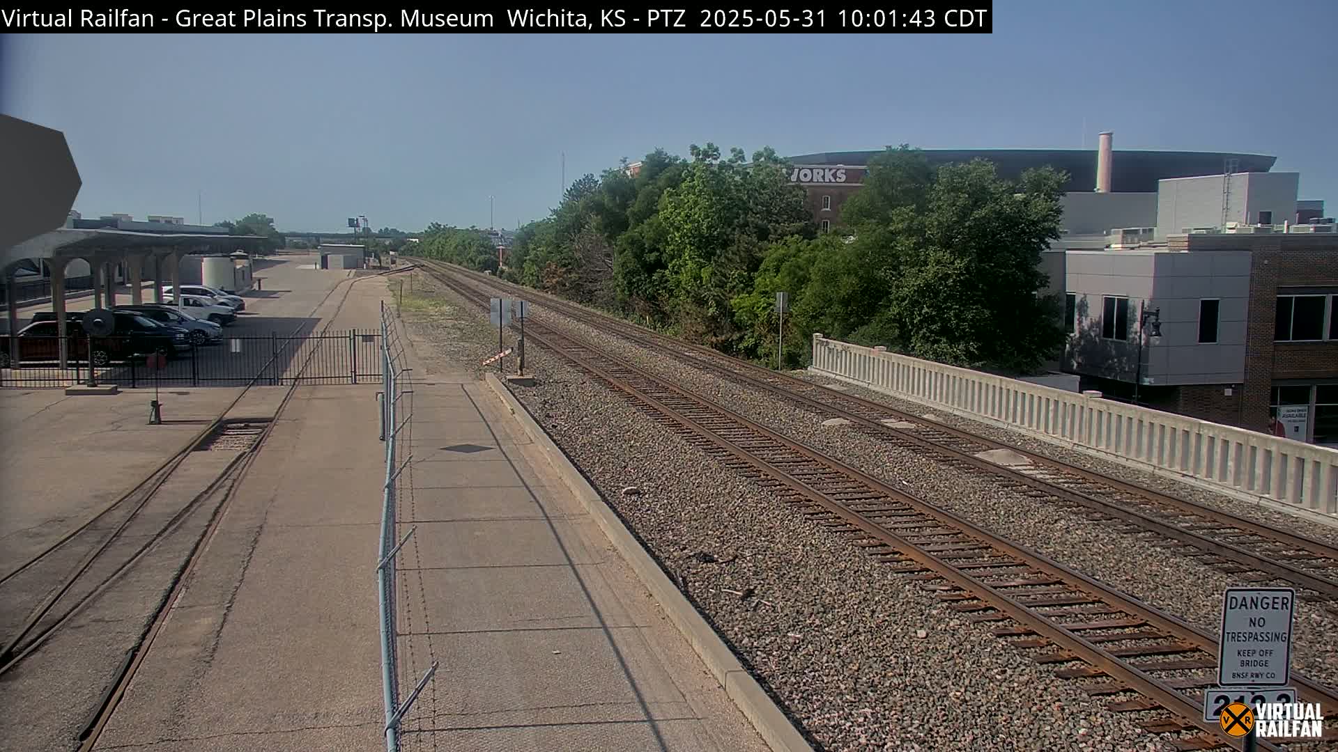 A clear sunny day view of railroad tracks next to a road, with parked cars visible to the left and buildings in the distance.