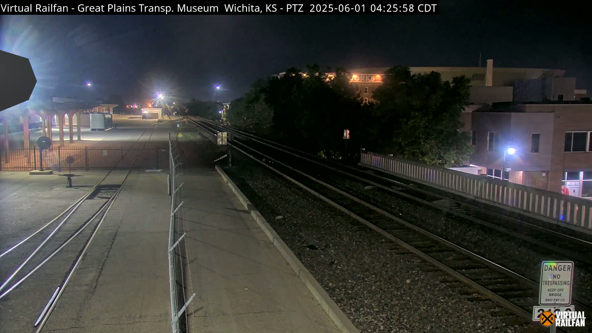 At night, train tracks extend into the distance past a fenced-in area with a building and some lights.