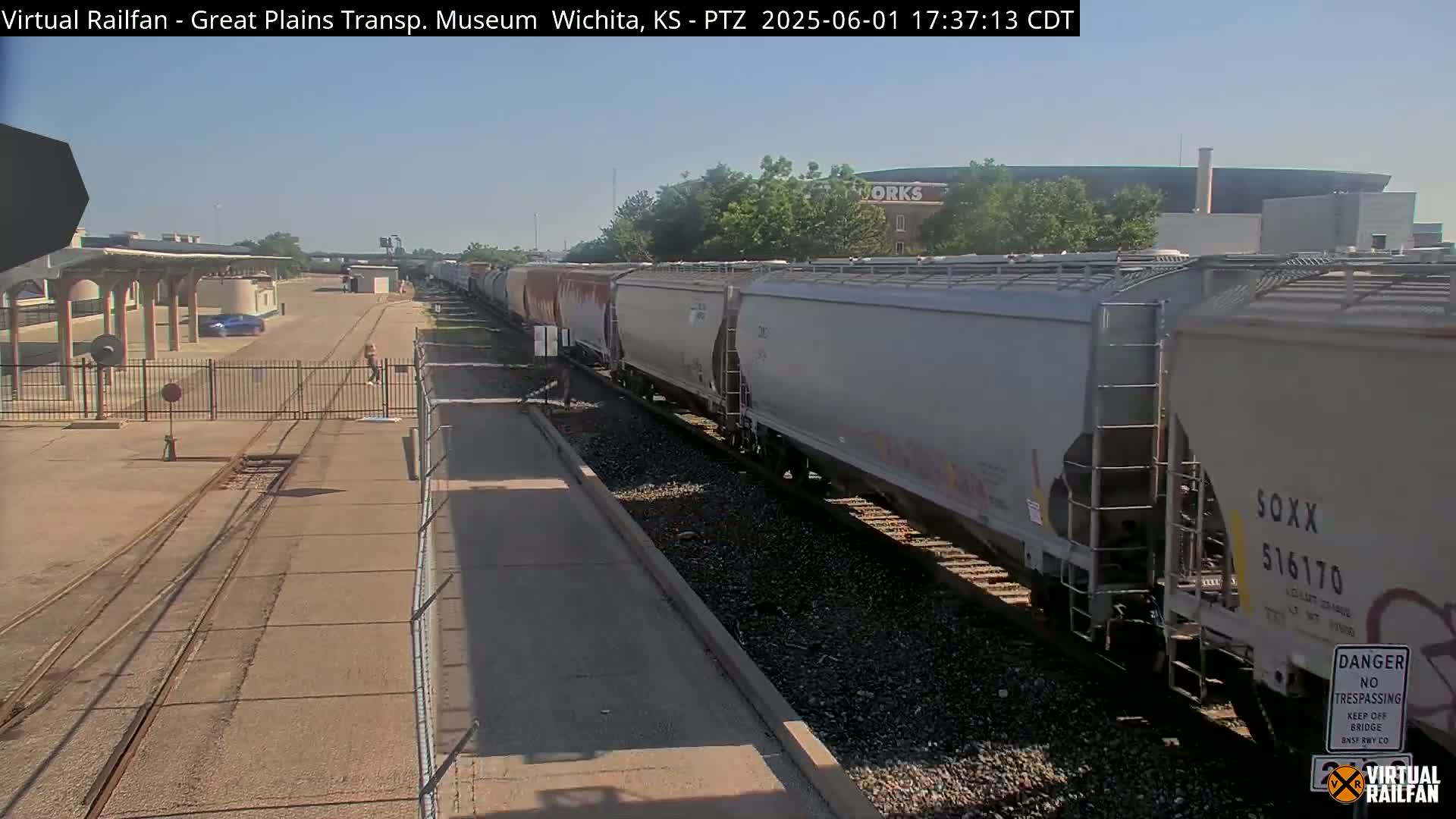 A long freight train of light gray covered hopper cars passes by a rail yard on a sunny day.