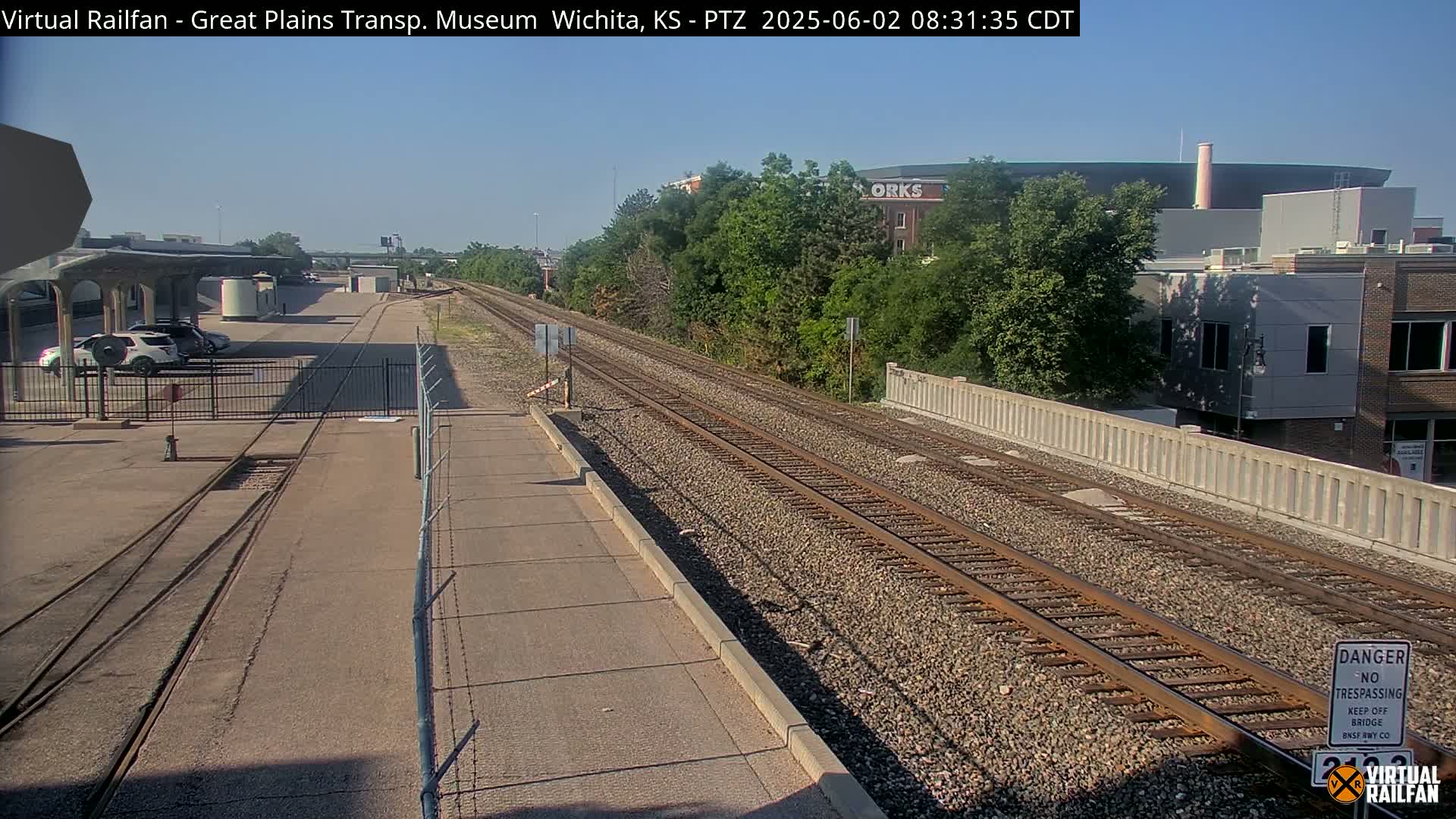 A sunny day view of railroad tracks extending into the distance, past a chain-link fence and a parking lot with cars, toward buildings and trees.