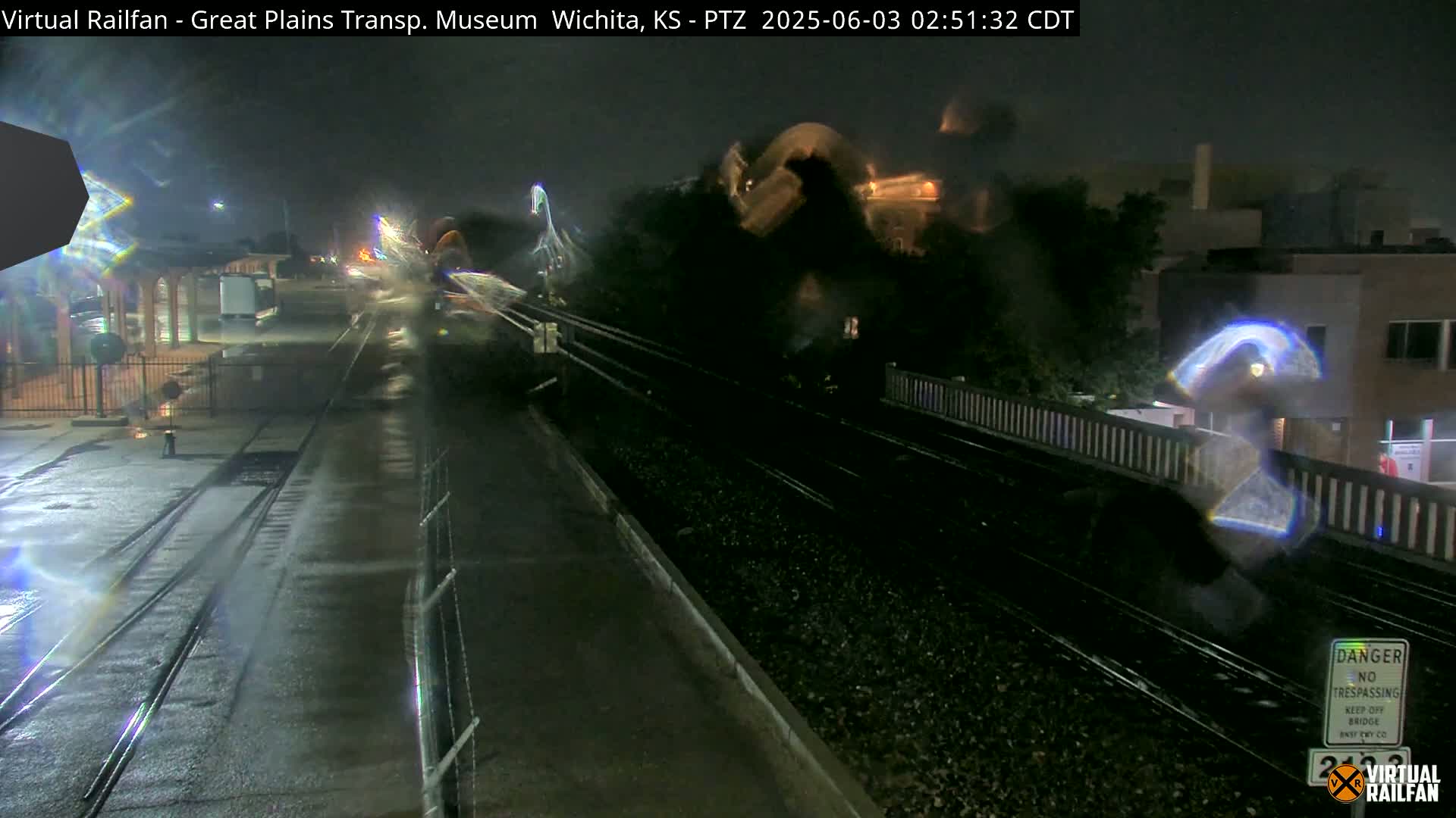 A nighttime view of rain-slicked train tracks and a nearby building, with dark, hazy conditions in the air.