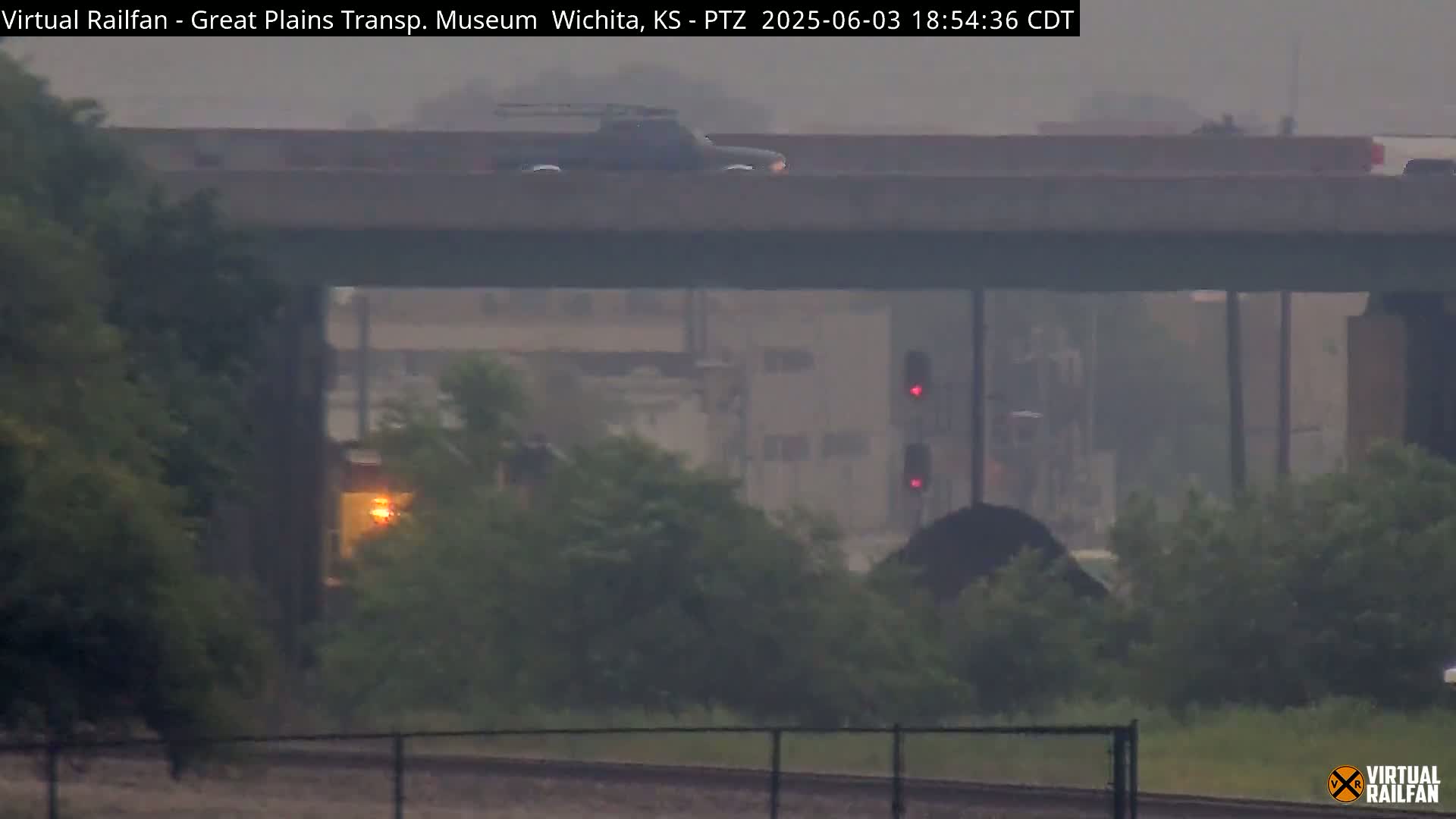 A dark-colored pickup truck drives across an overpass in hazy, misty conditions, with a train partially obscured by vegetation and mist below.