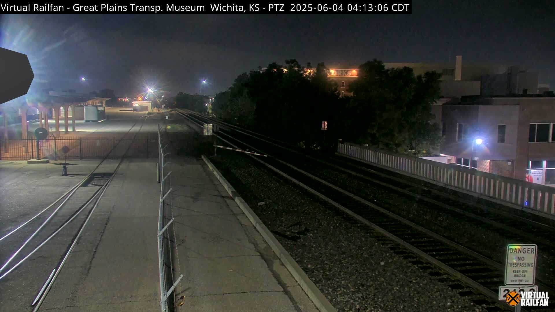 At night, train tracks extend into the distance, passing near buildings under a dark sky.