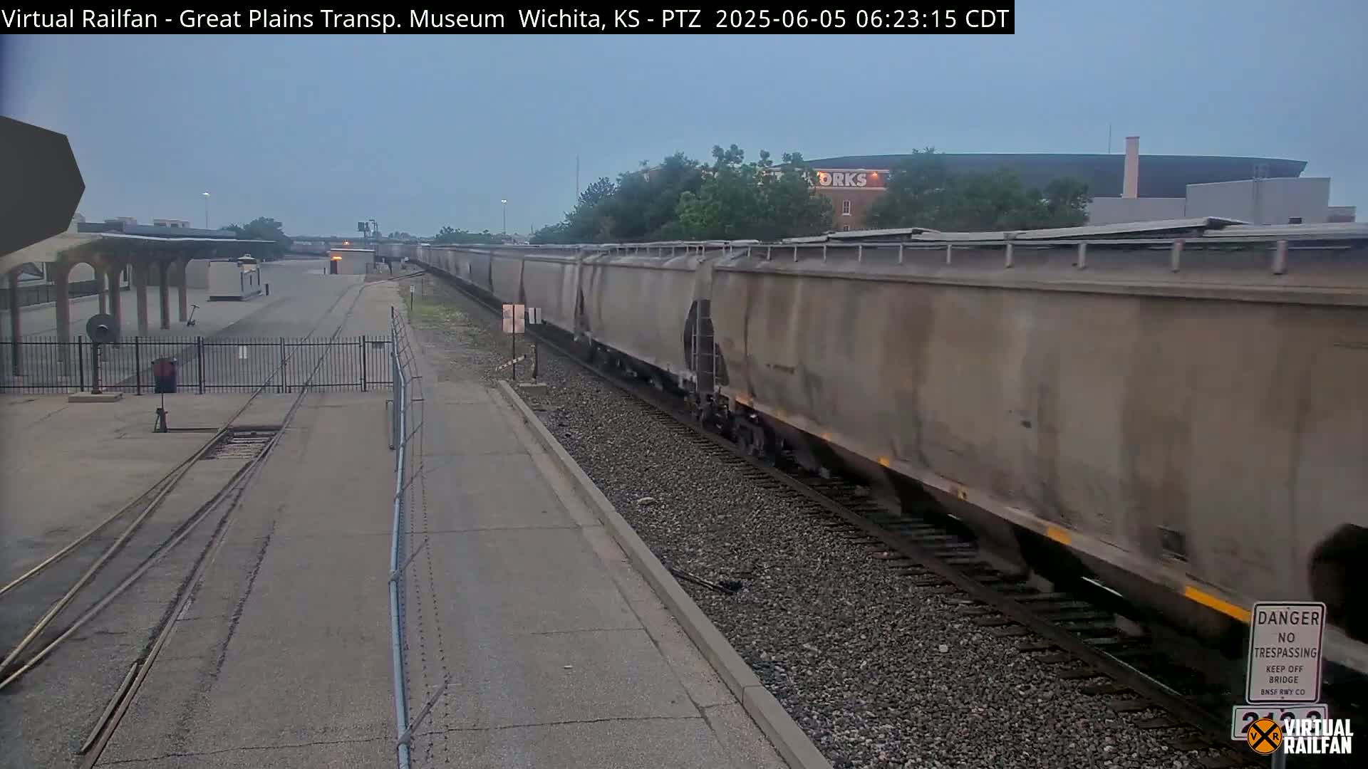 A long freight train passes by a railway station under an overcast sky.