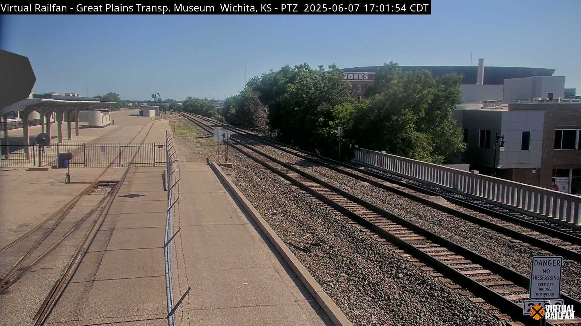 A sunny day reveals a railway scene with multiple tracks leading towards a bridge and buildings beyond, bordered by a fence.