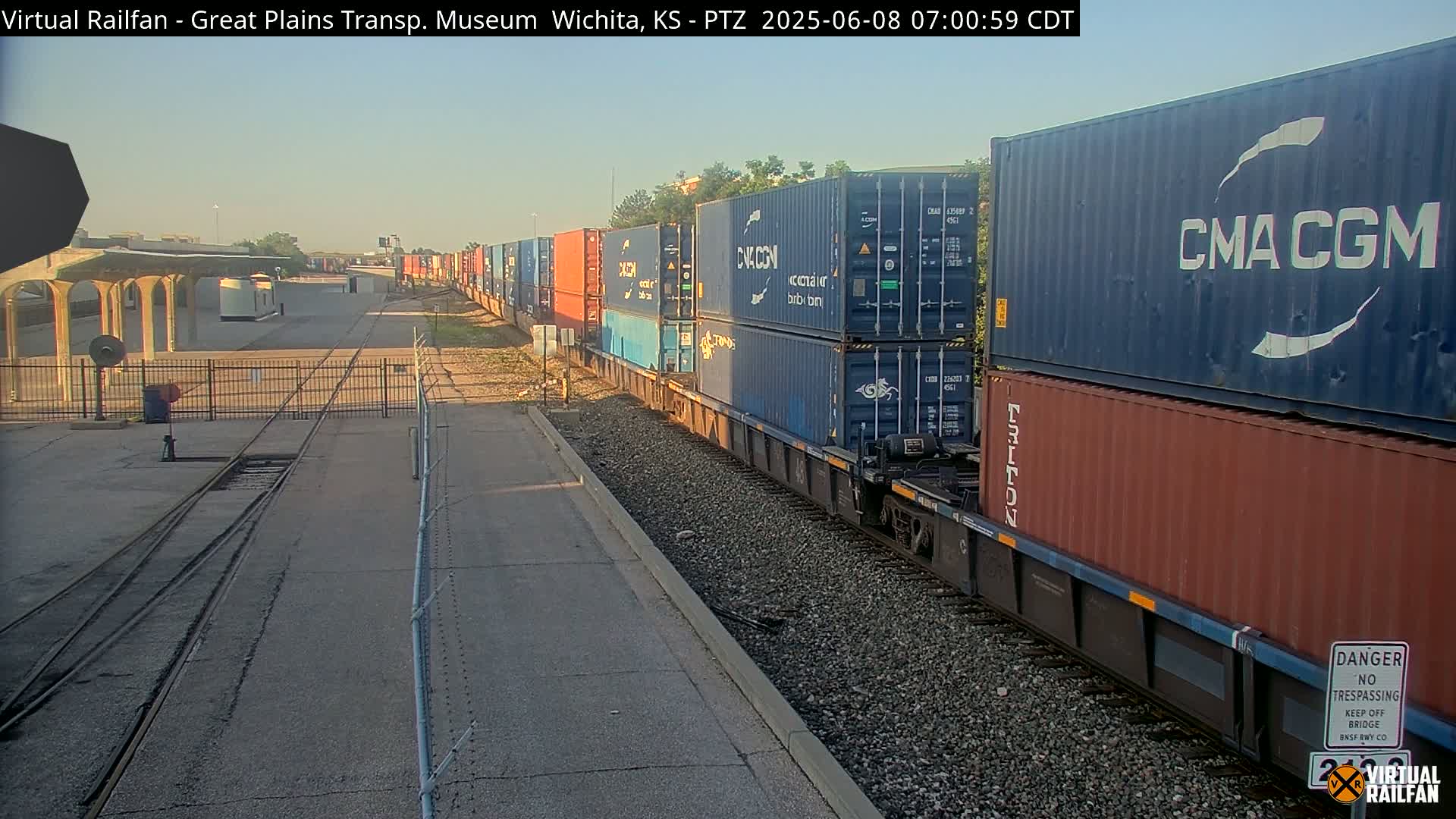 A long freight train carrying stacked shipping containers moves along a railway track under a clear, bright sky.