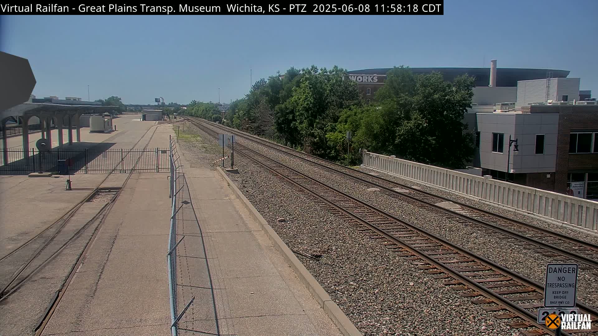 A sunny view of railroad tracks extending from a paved area with a chain-link fence, passing alongside a low wall and greenery towards buildings in the distance.