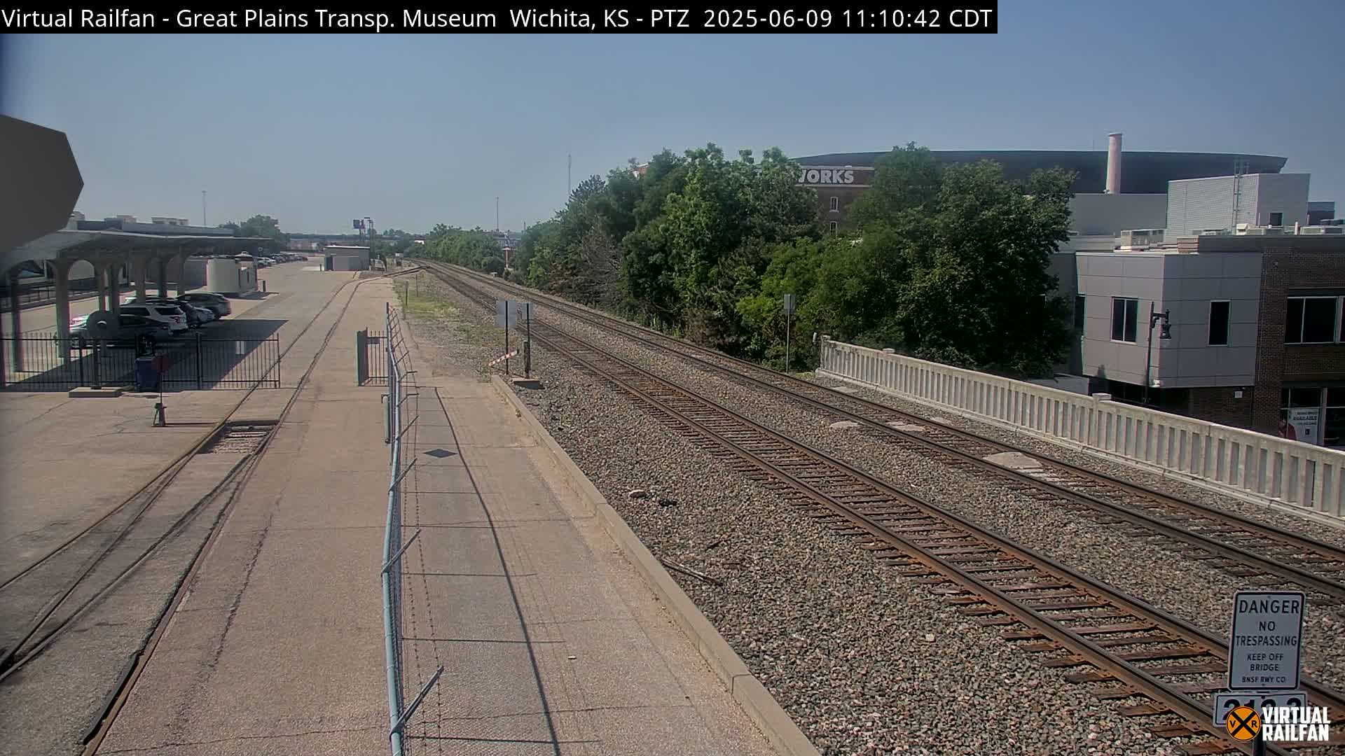 A sunny view of railroad tracks extending into the distance, passing by a parking lot, some buildings, and lush green trees.