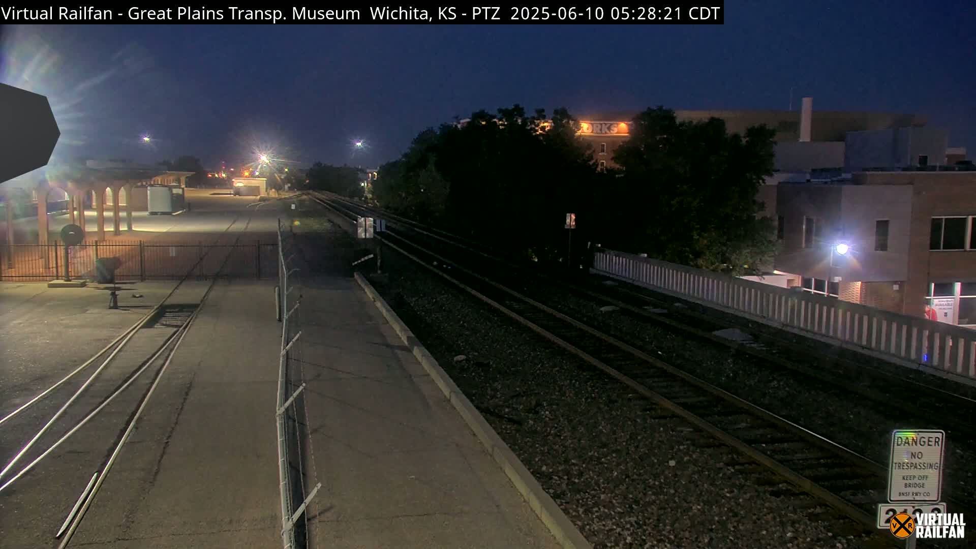 At night, train tracks extend into the distance past a building and a fenced-in area with a small structure.