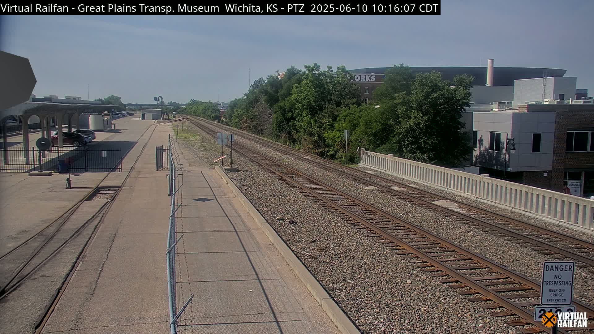 A sunny day reveals a train track alongside a paved road, with a parking lot and buildings visible in the distance.