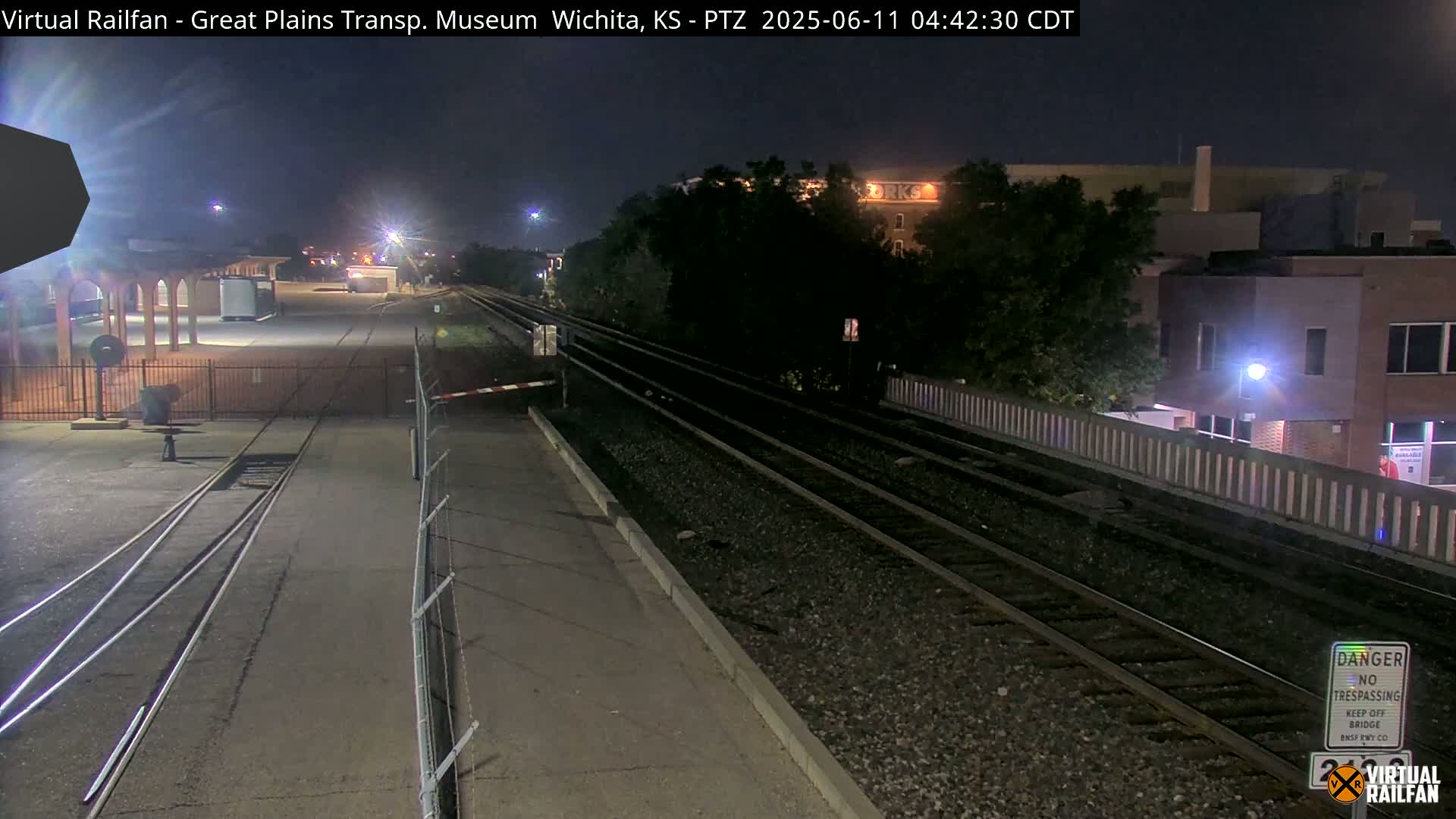 A nighttime view of train tracks and a station platform, with buildings and trees visible in the background under dark skies.