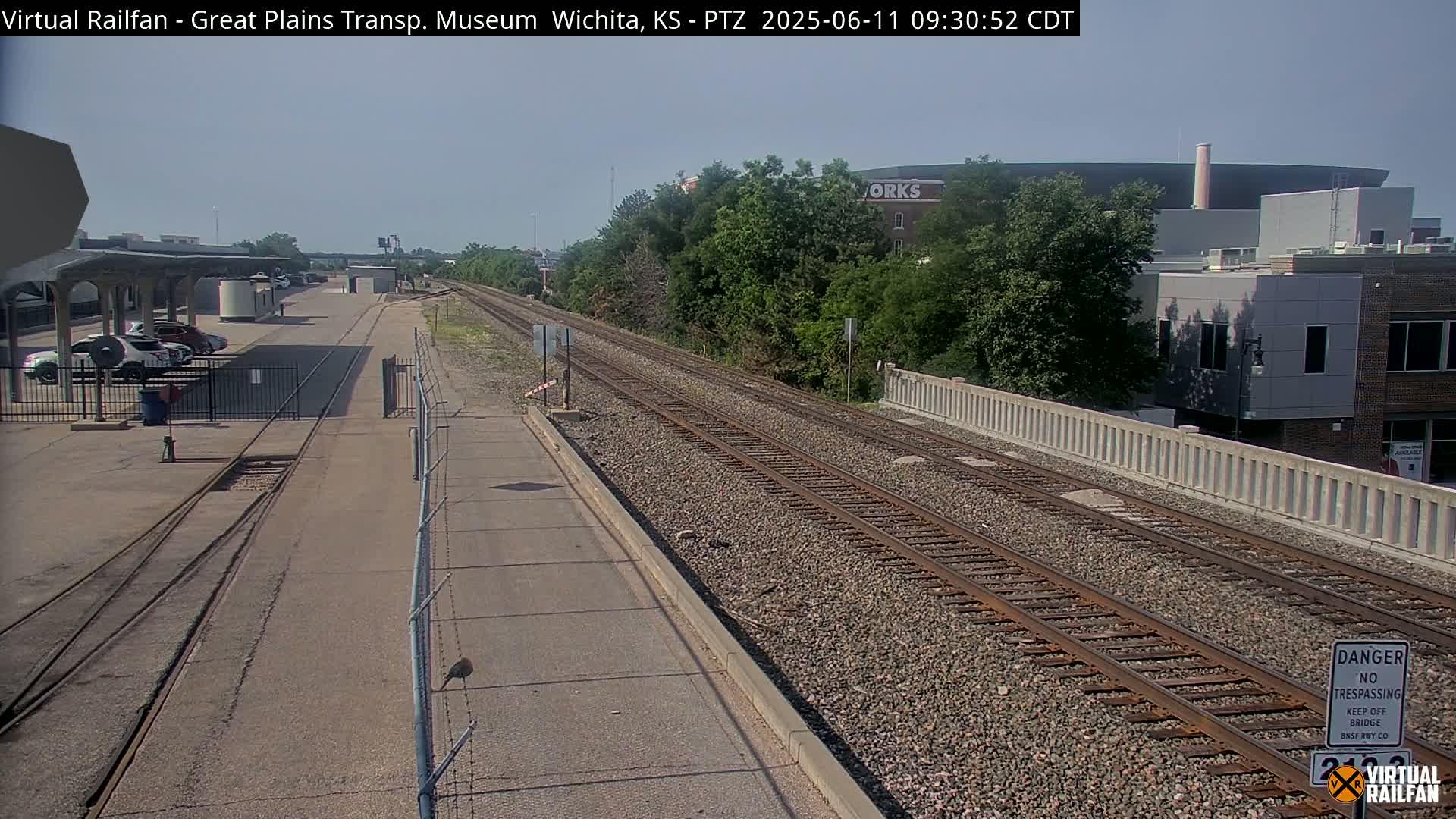 A sunny day at a train station shows multiple train tracks, a paved road, and buildings in the background.