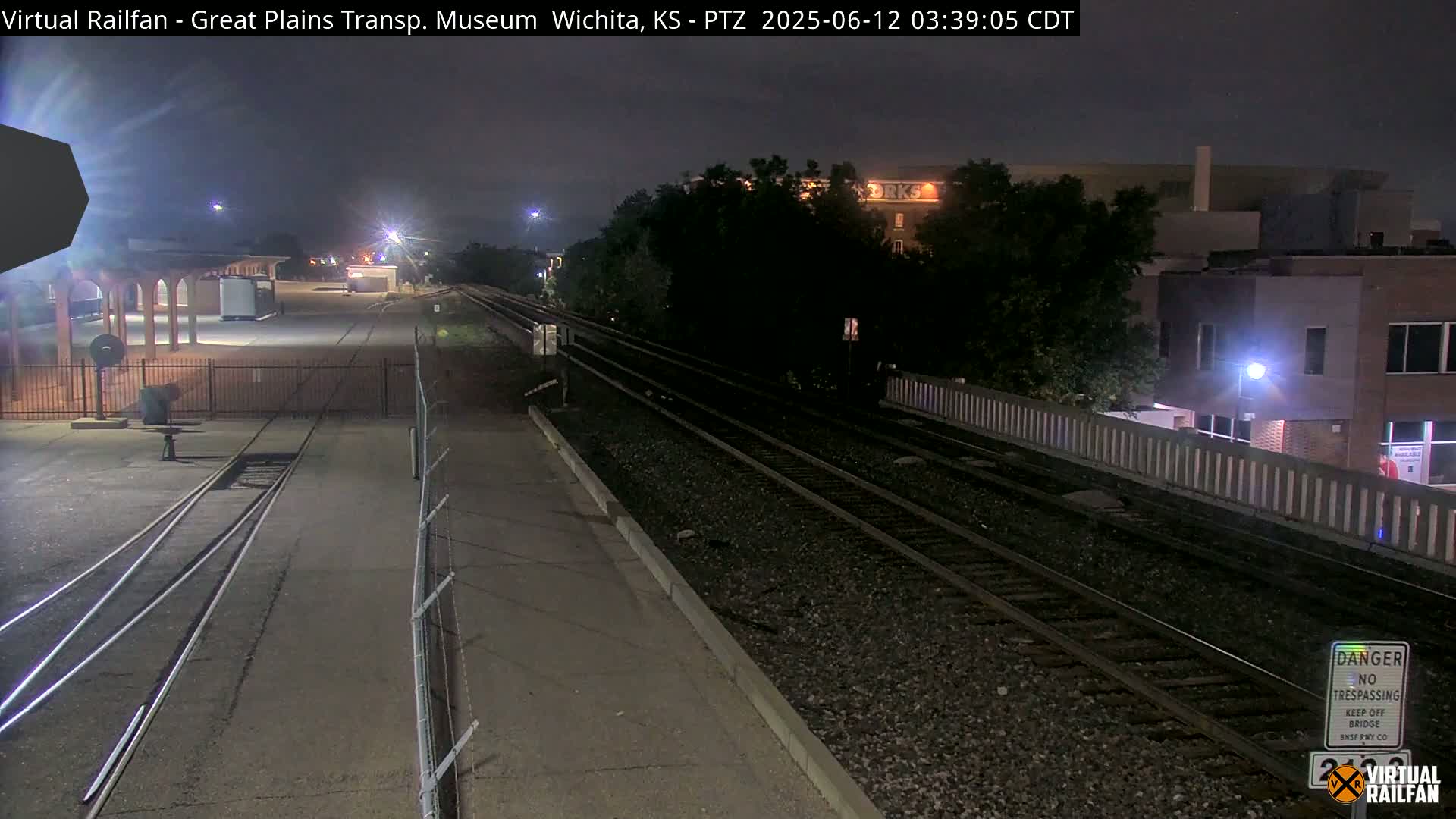 At night, railroad tracks extend into the distance past a low brick wall and a building, under a dark sky.