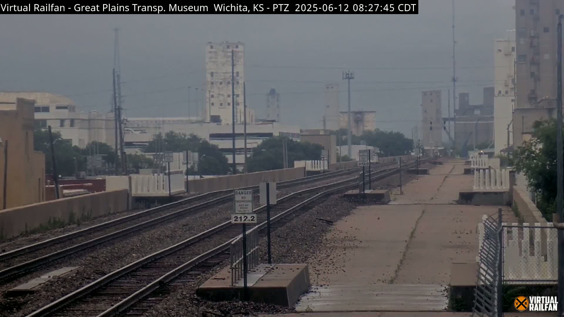A hazy view of railroad tracks stretches into the distance, passing between industrial buildings and low-lying vegetation under an overcast sky.