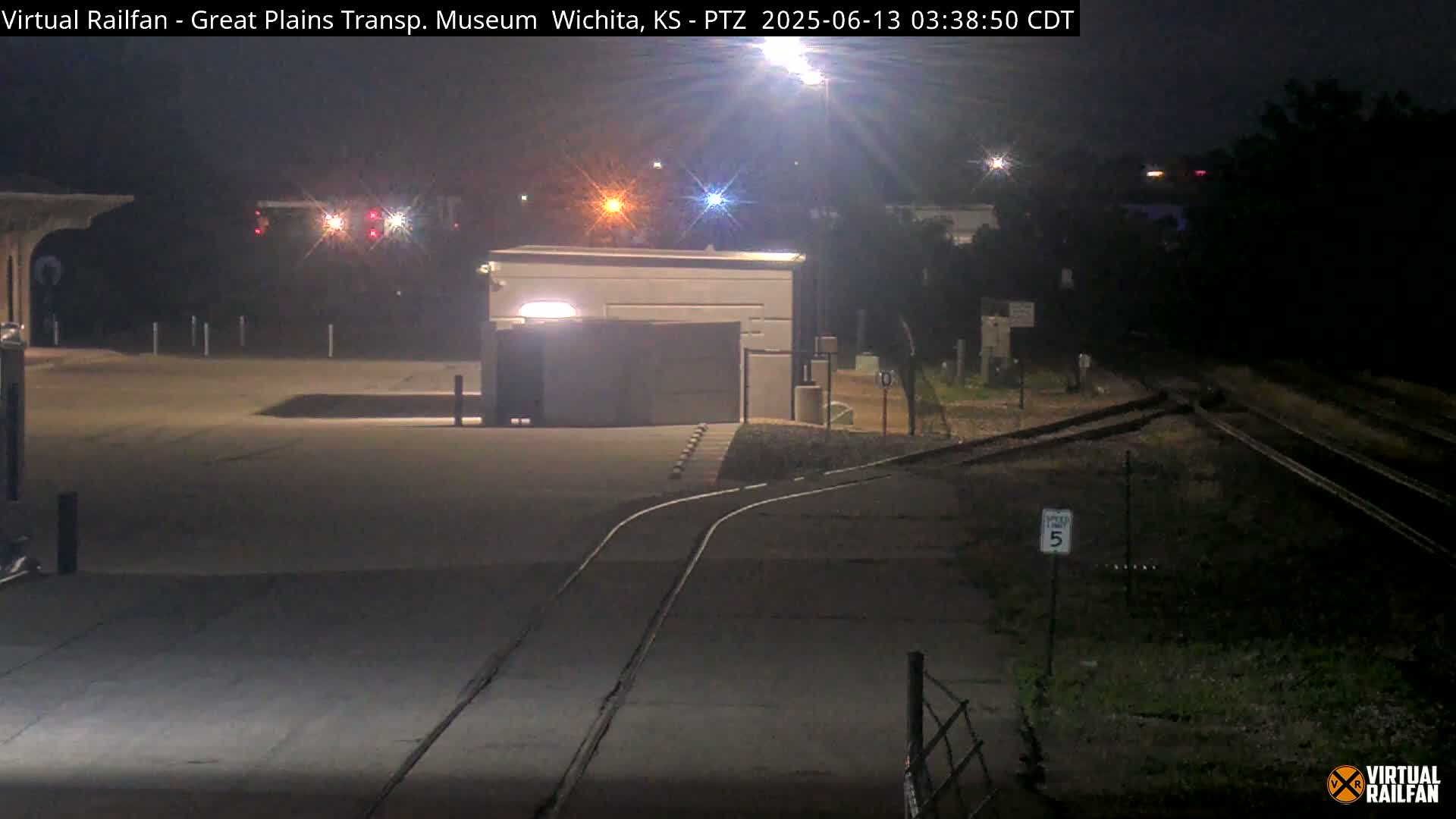 A nighttime view of a railway crossing near a building with a closed gate, under artificial light.